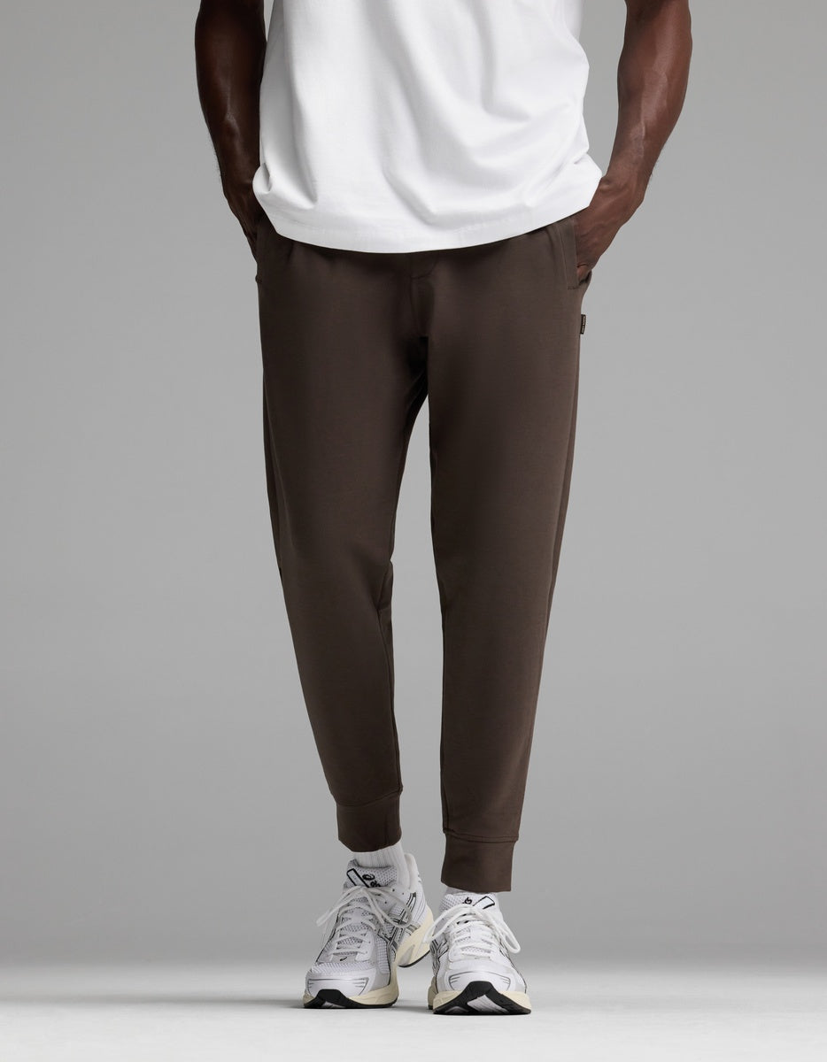 A person is shown from the waist down wearing Cozy Earth’s Men’s StretchTech Jogger in dark brown, paired with a white T-shirt and white sneakers, standing against a plain gray background.