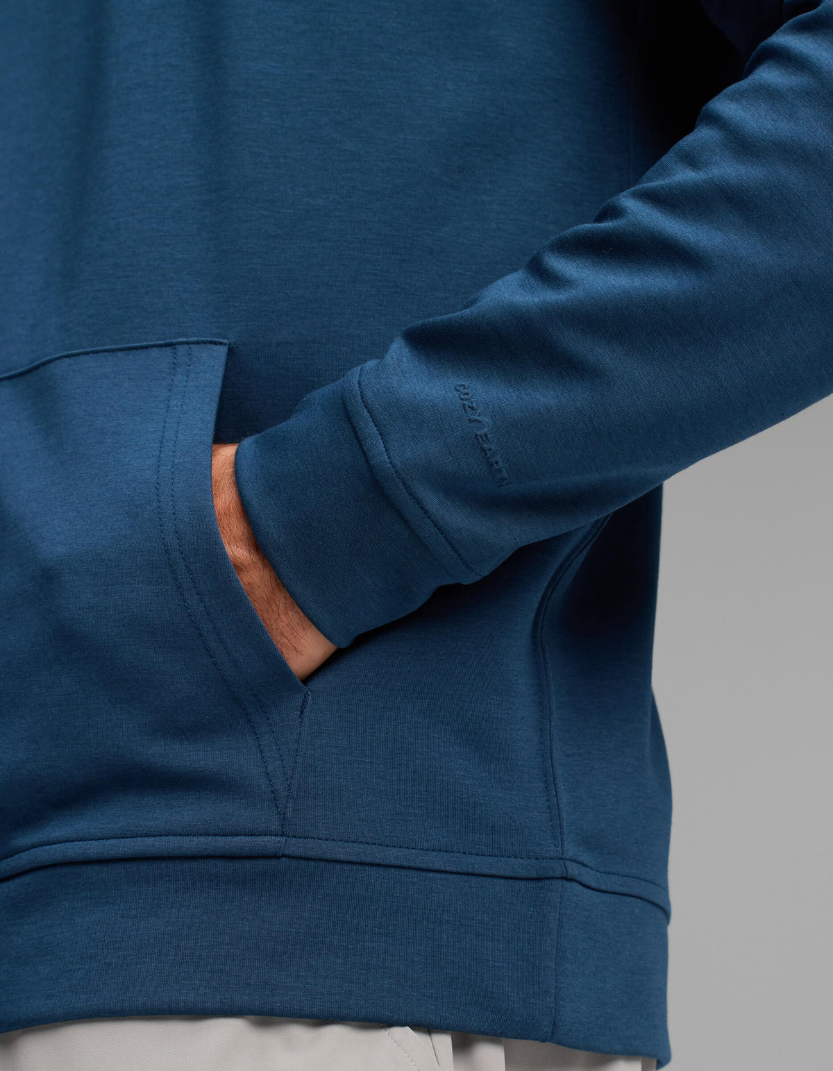 A man wearing the Cozy Earth Men's Versa Hoodie in blue, with one hand in the front pocket, stands against a plain gray background.