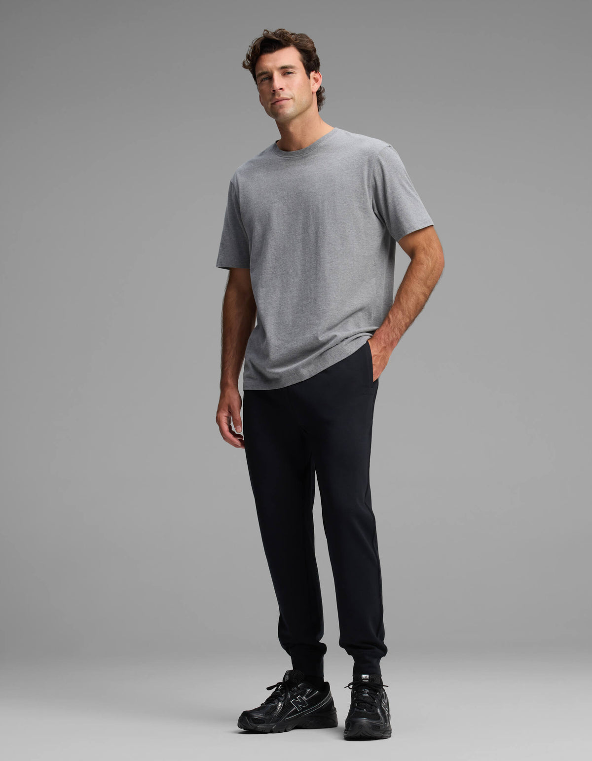 A man stands against a plain gray background, wearing a gray short-sleeve t-shirt, black sneakers, and the Cozy Earth Men's Versa Jogger in black, with one hand in his pocket.
