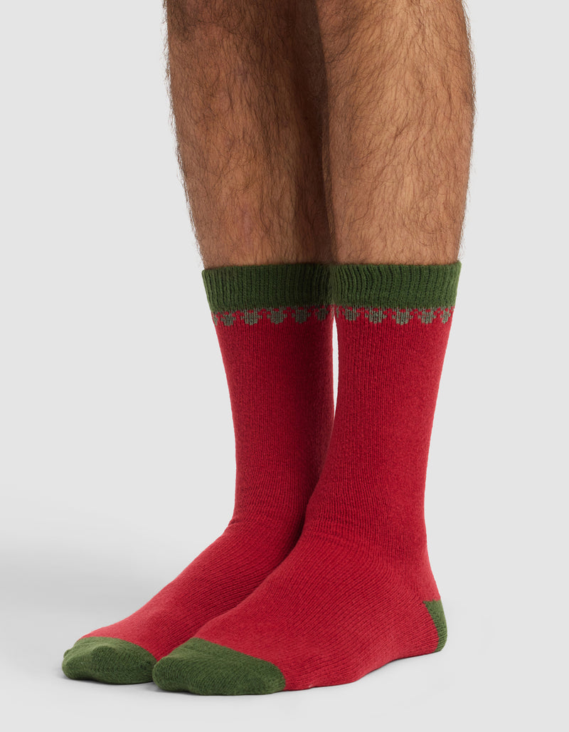A person wears The Mistletoe Plush Lounge Sock by Cozy Earth—red socks with green heels, toes, and cuffs plus a small green and gray pattern near the top. The plain background is light-colored.