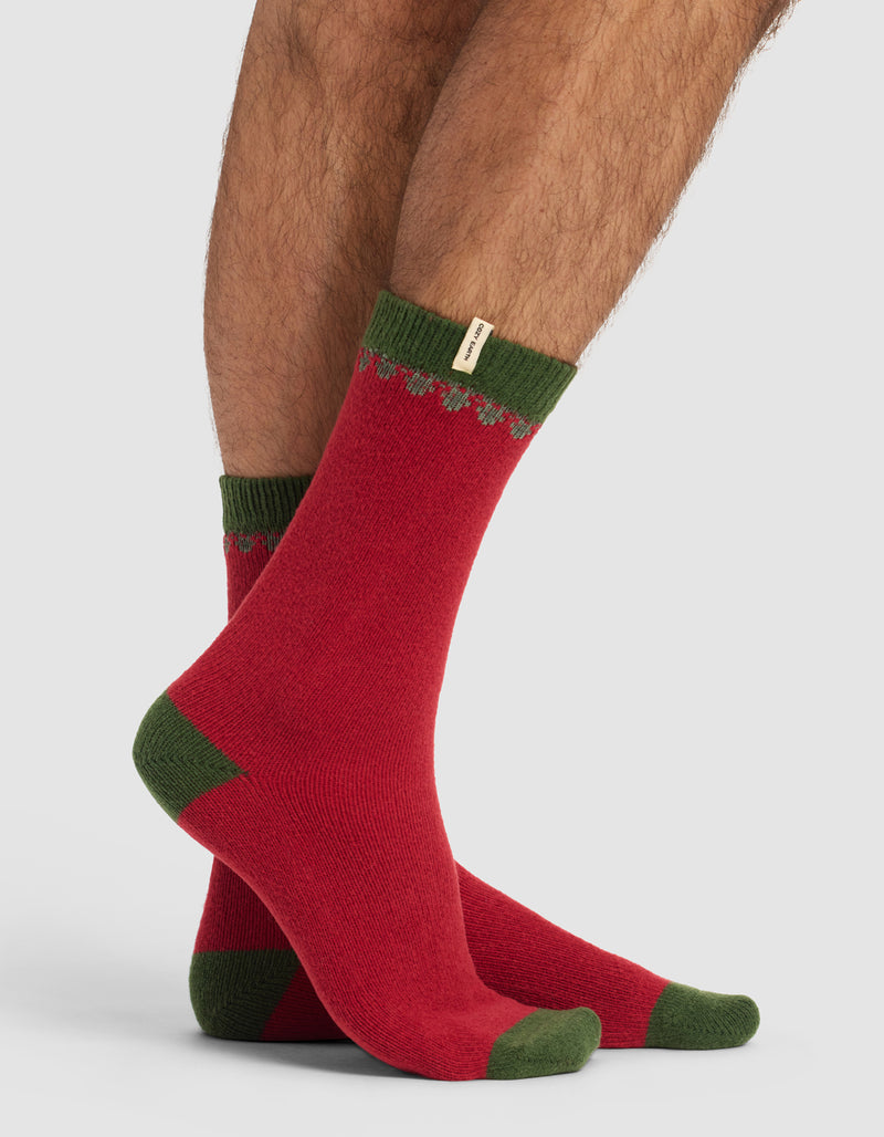 A person wearing The Mistletoe Plush Lounge Sock by Cozy Earth—red with green toes, heels, and cuffs, featuring a small decorative tag and a subtle zigzag pattern near the top—stands against a plain light gray background.