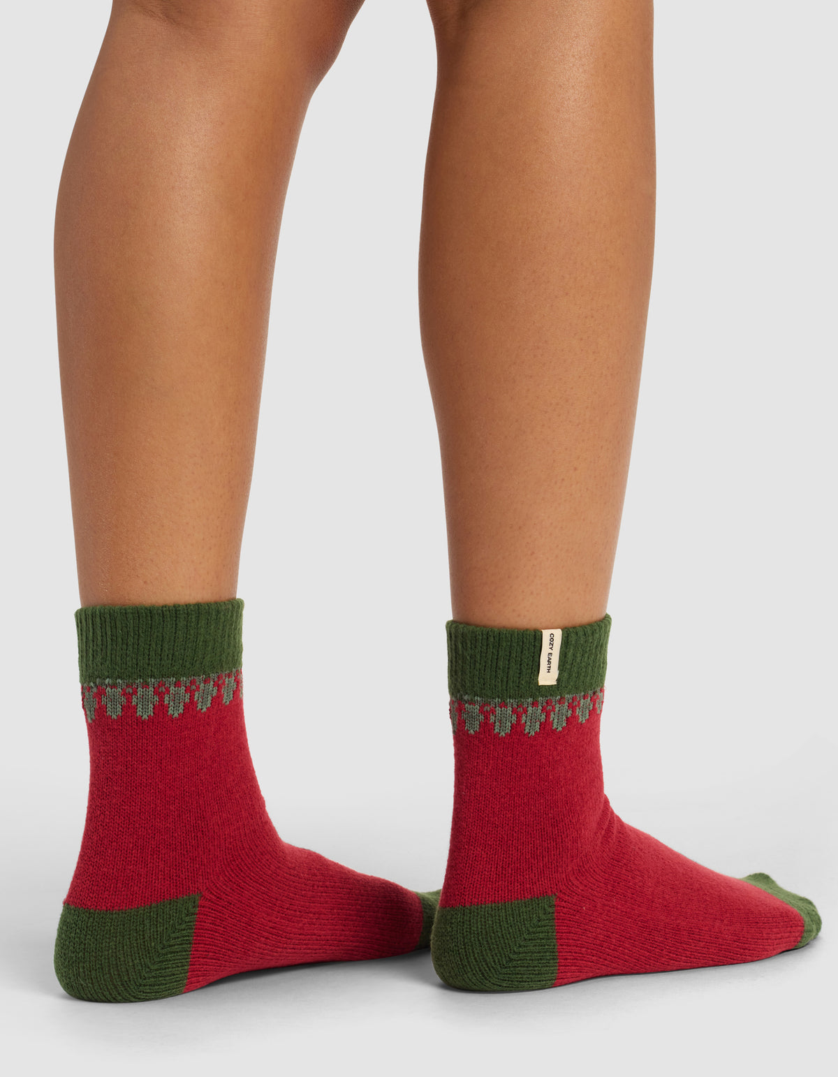 A close-up of a person’s lower legs in Cozy Earth’s The Mistletoe Plush Modern Crew Sock—red with green cuffs, heels, and toes, plus a small gray pattern near the cuffs—against a plain background.