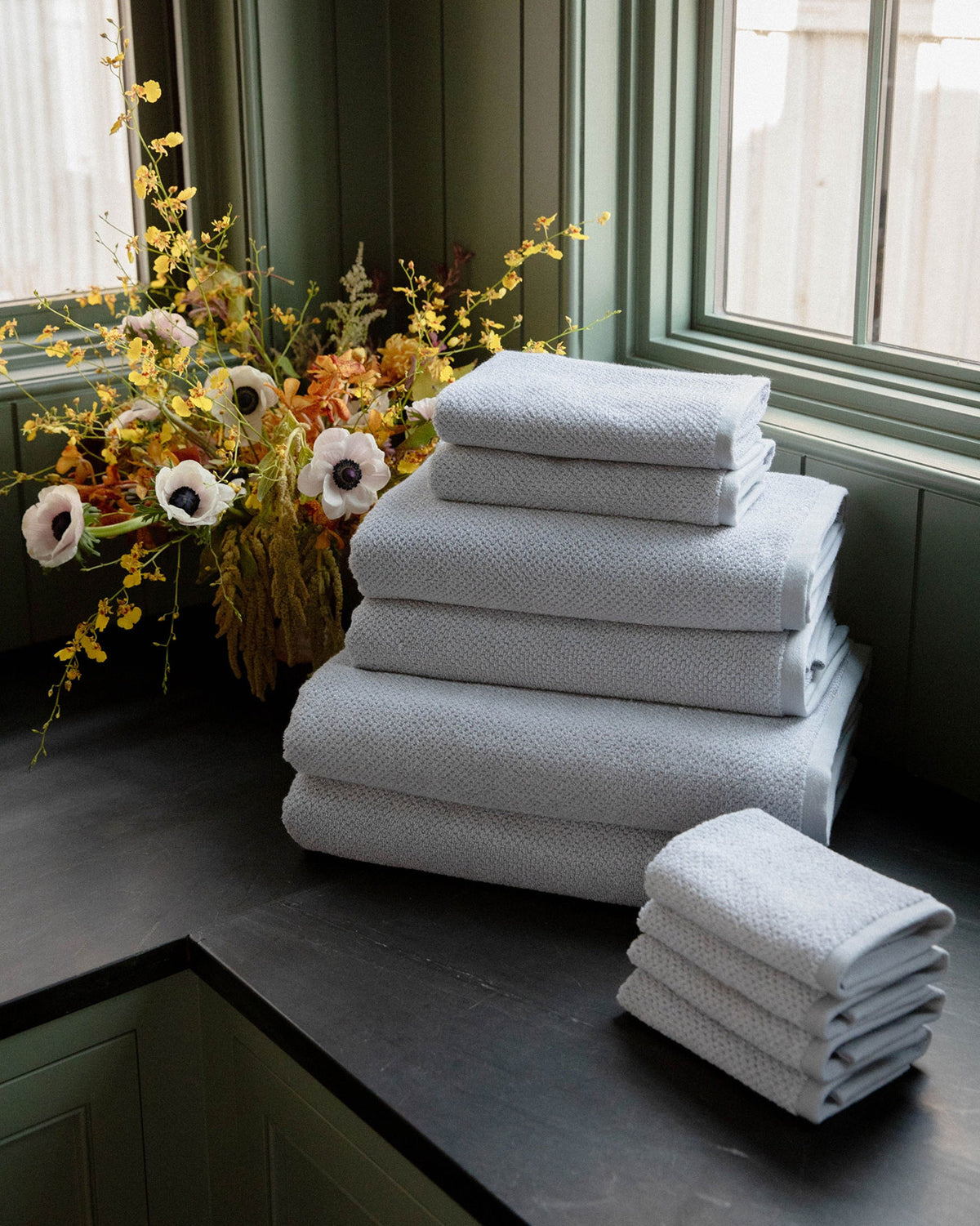 The Complete Nantucket Bath Bundle by Cozy Earth is neatly stacked on a dark countertop near a window, with a vase of yellow and white flowers in the background.