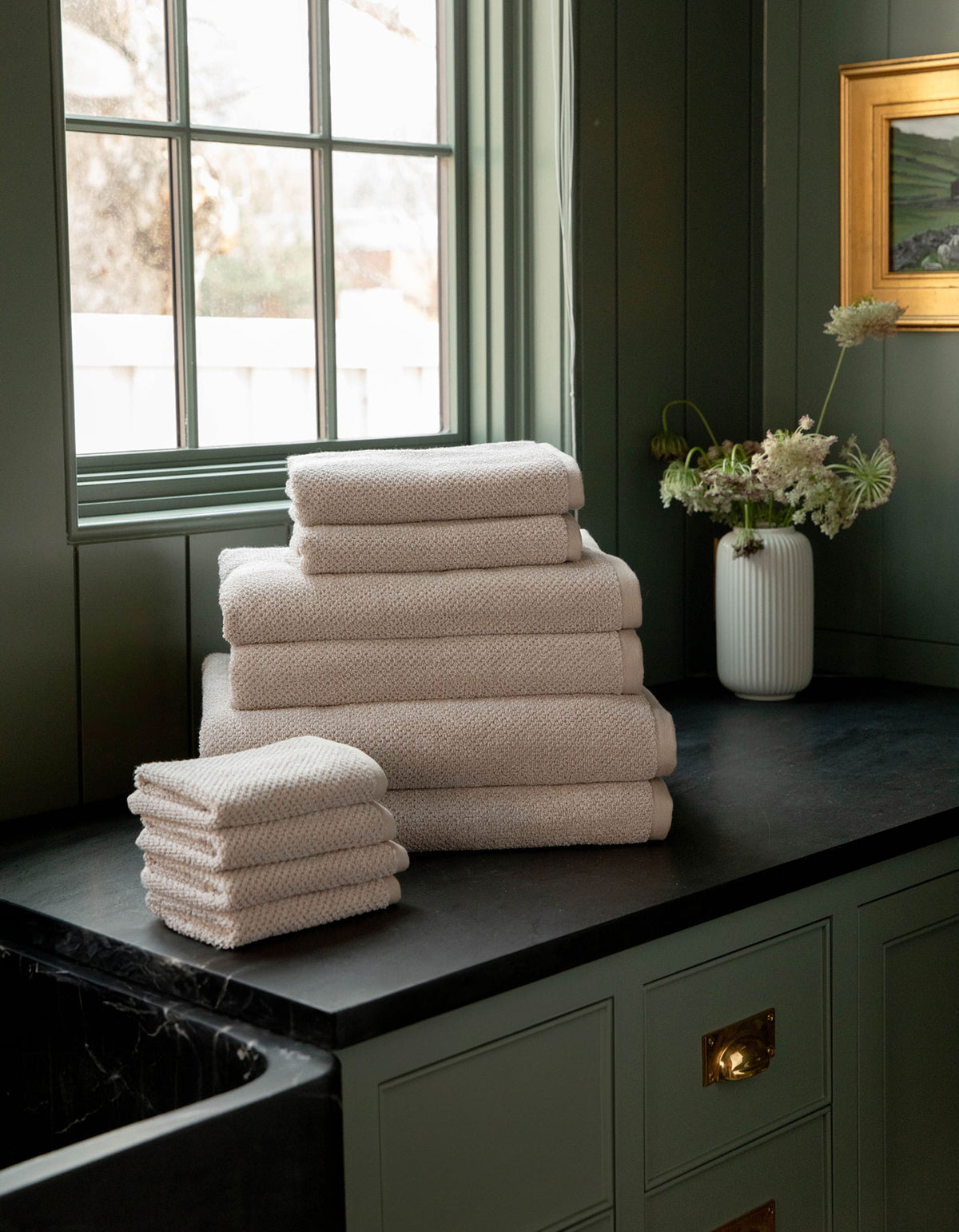 The Complete Nantucket Bath Bundle by Cozy Earth, in beige, is neatly stacked on a black countertop near a window with a vase of flowers and pale green cabinetry in the background.
