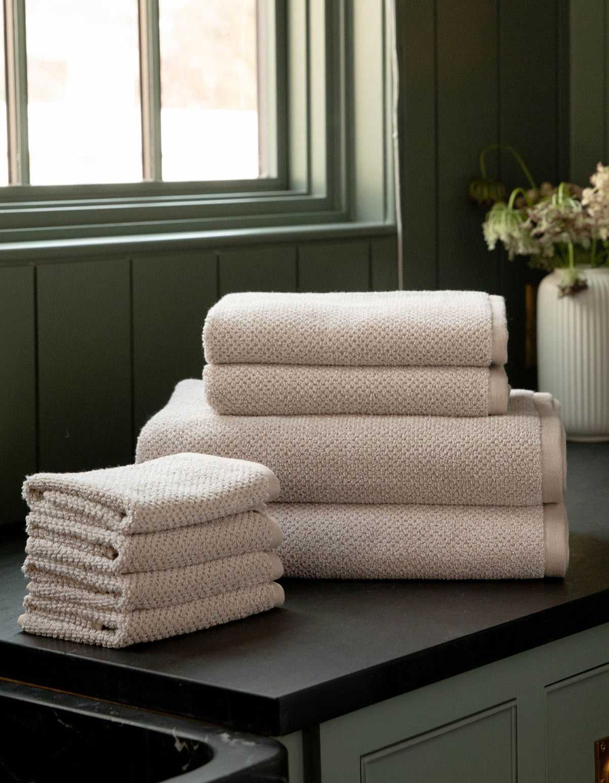 The Cozy Earth Nantucket Bath Towel Set, featuring hand and bath towels in beige, is neatly stacked on a dark countertop near a window with green paneling. A white vase with flowers appears in the background.