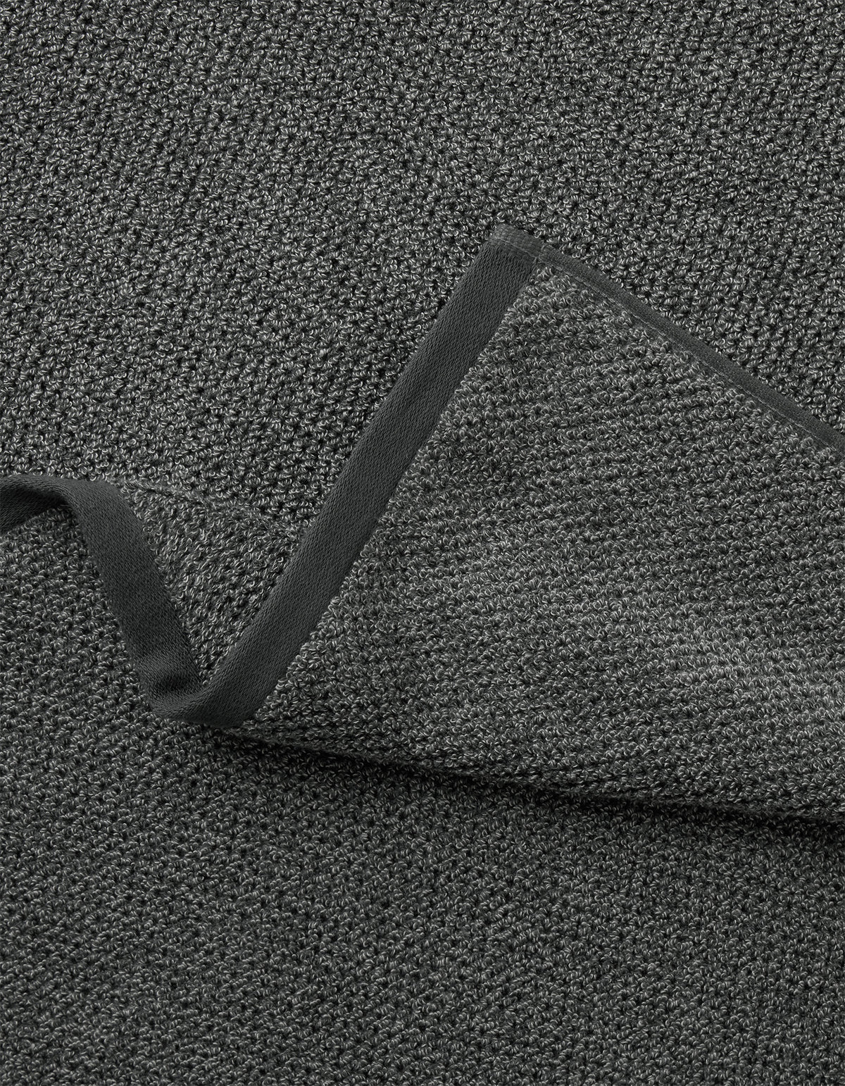 A close-up of Cozy Earth's Nantucket Bath Towel Set shows its textured dark gray fabric with a folded corner, revealing the same woven material on both sides and a smooth, dark border along the edge.