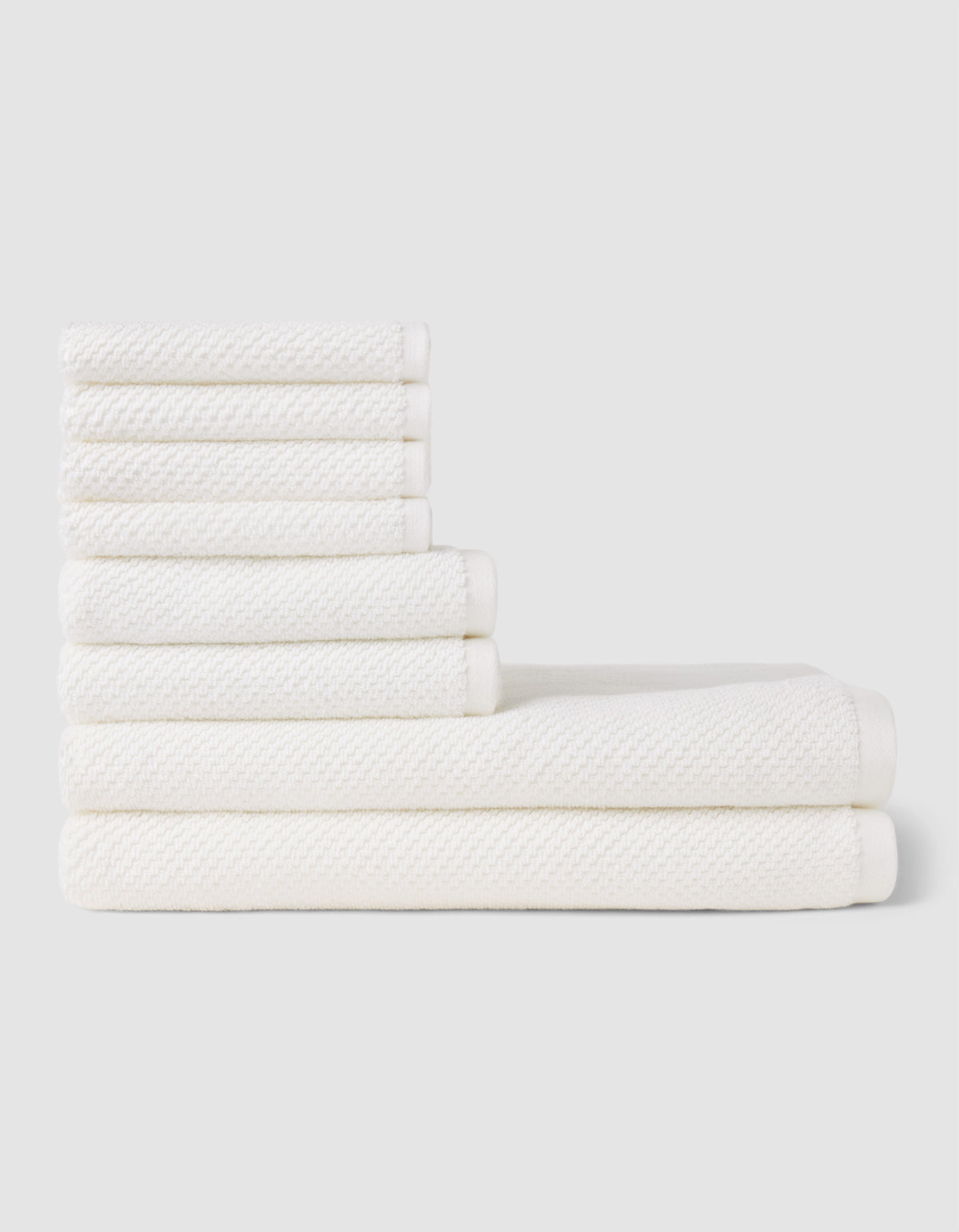 The Cozy Earth Nantucket Bath Towel Set features seven white textured towels—three large and four small—neatly stacked in two rows on a light gray background, showcasing their soft, plush feel.|Color:White