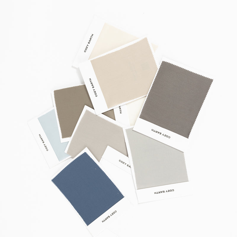 A variety of "Cozy Earth" Bamboo Sheet Swatches are laid out on a white surface, showcasing an array of colors including driftwood, oat, walnut, charcoal, light grey, dove grey, pacific blue, shore, creme, and white