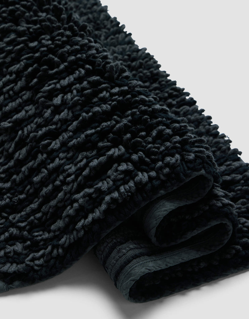 A close-up of the Plush Loop Bath Mat by Cozy Earth shows its dark, textured fabric with a thick, looped pile and soft, plush folds against a plain light background.