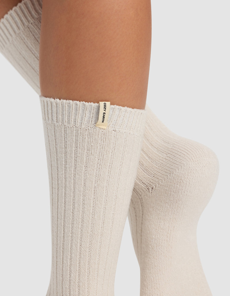 Close-up of crossed legs wearing thick, ribbed, cream-colored socks from the Plush Lounge Calf Sock 3-Pack by Cozy Earth. A small cream tag with black “COZY EARTH” text is on the ribbed cuff. Background is plain white.