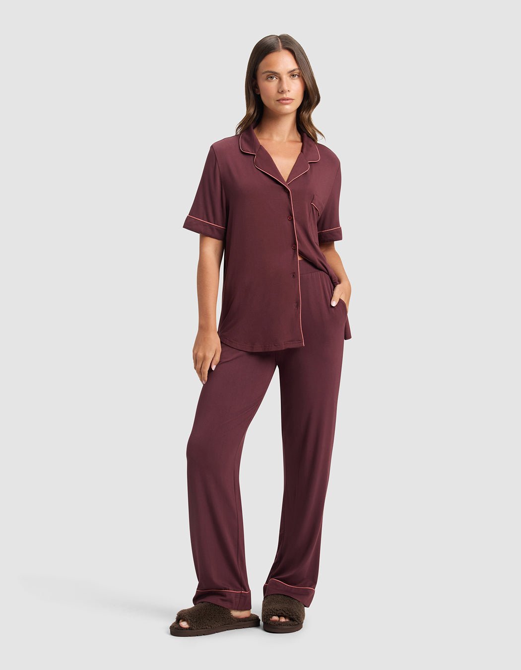 A woman stands facing forward, wearing a maroon pajama set with short sleeves and long pants. She has her left hand in her pocket and is wearing Cozy Earth Puffy Sheep Slides. The background is plain light gray.|Color:Espresso