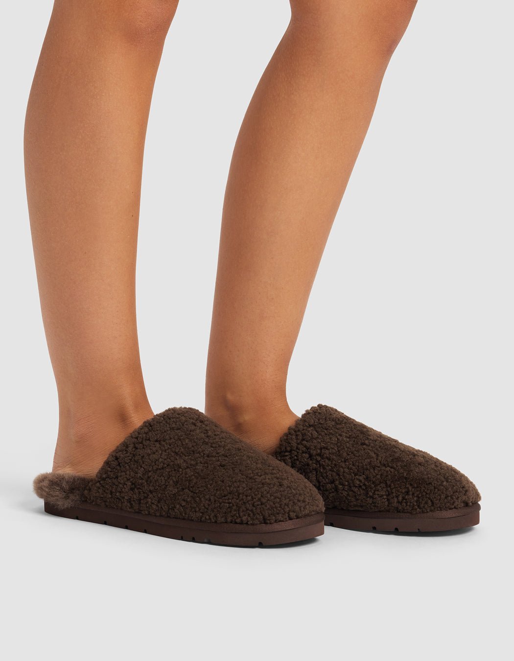 A person wears Cozy Earth Puffy Sheep Slippers—dark brown, fuzzy, slip-on flats. Only their lower legs and feet are visible against a light gray background.|Color:Espresso