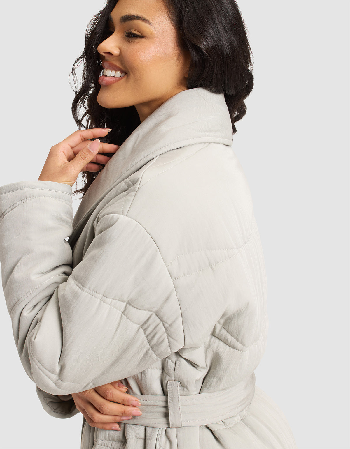 A woman with wavy dark hair smiles in a Cozy Earth Women's Quilted House Coat, light gray with a wide collar and belt, shown from the side against a plain light background. 
