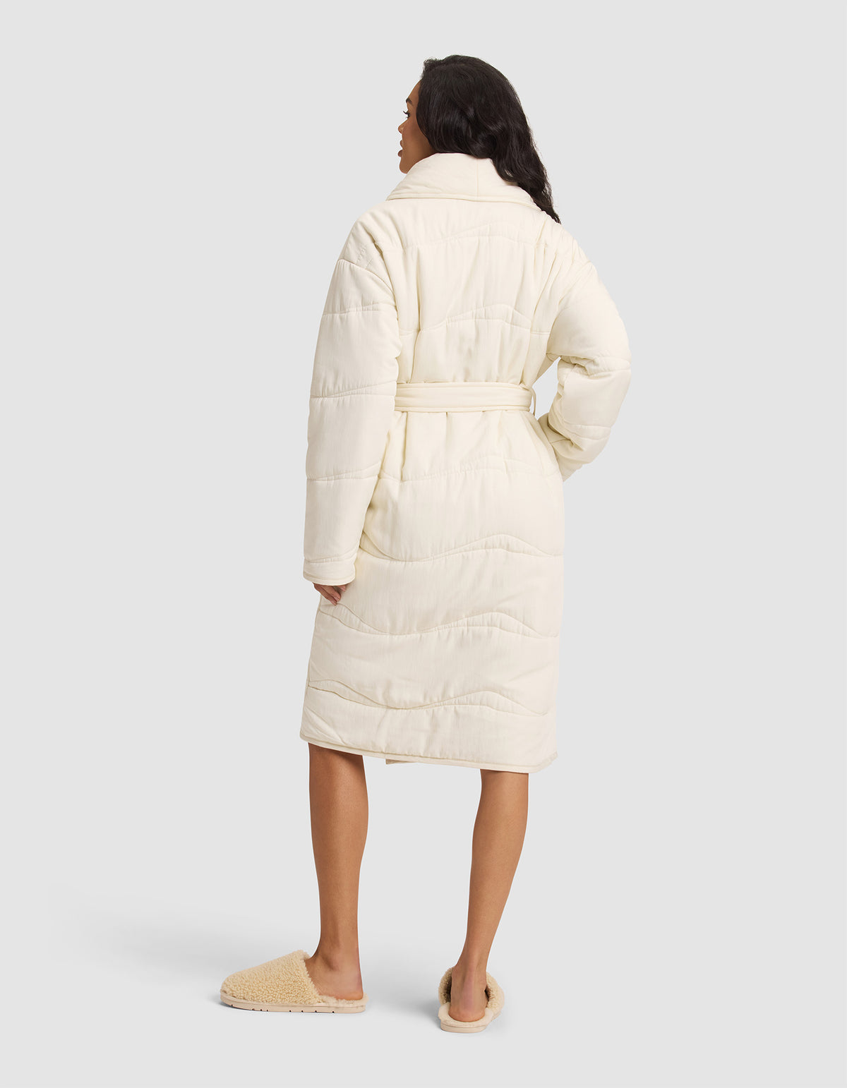 A woman faces away, wearing the Cozy Earth Women's Quilted House Coat in cream with a belt and tan slippers, set against a plain light gray background. 