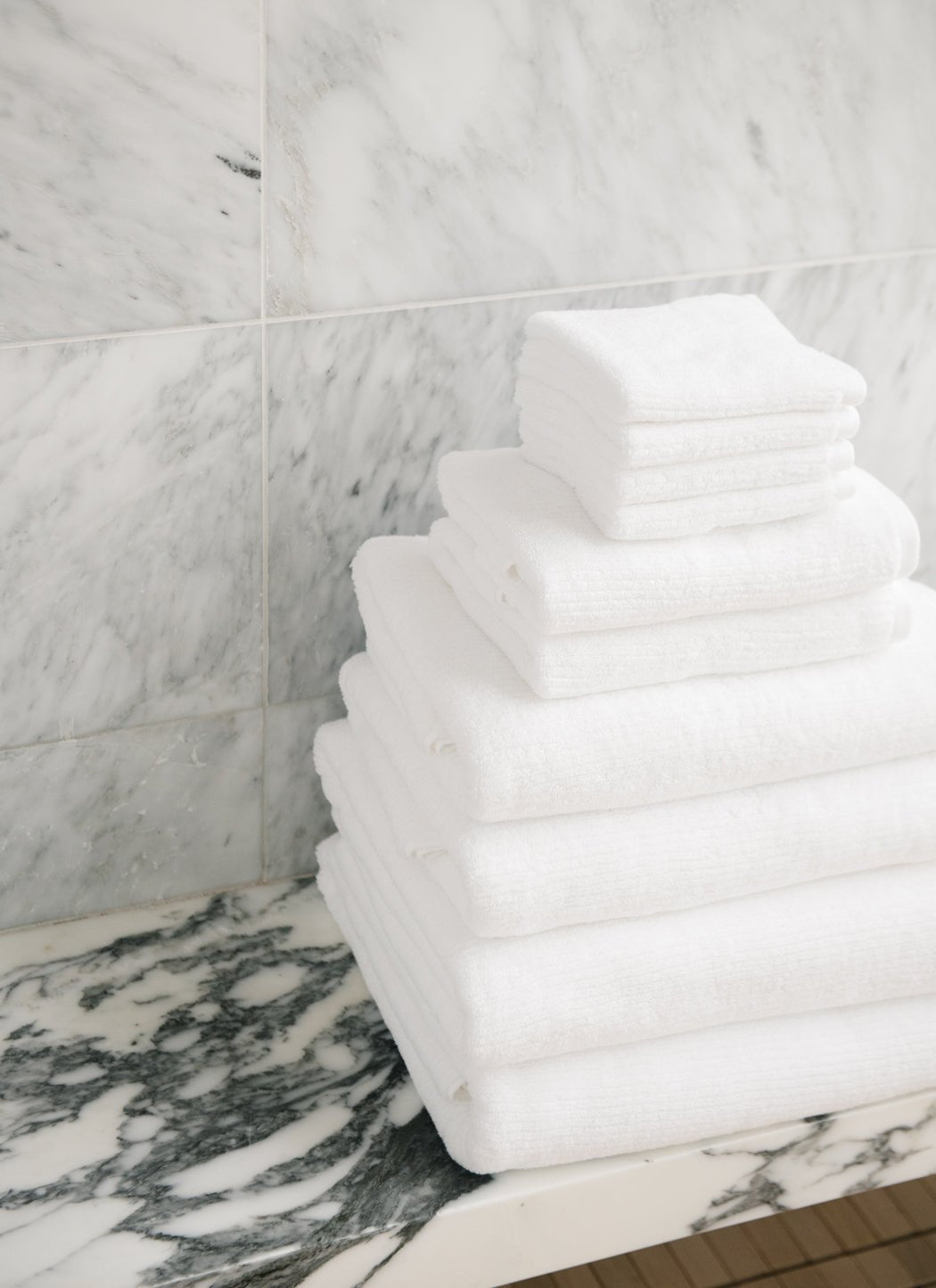 The Complete Ribbed Terry Bath Bundle by Cozy Earth is neatly stacked on a marble countertop, set against a gray and white marble wall.