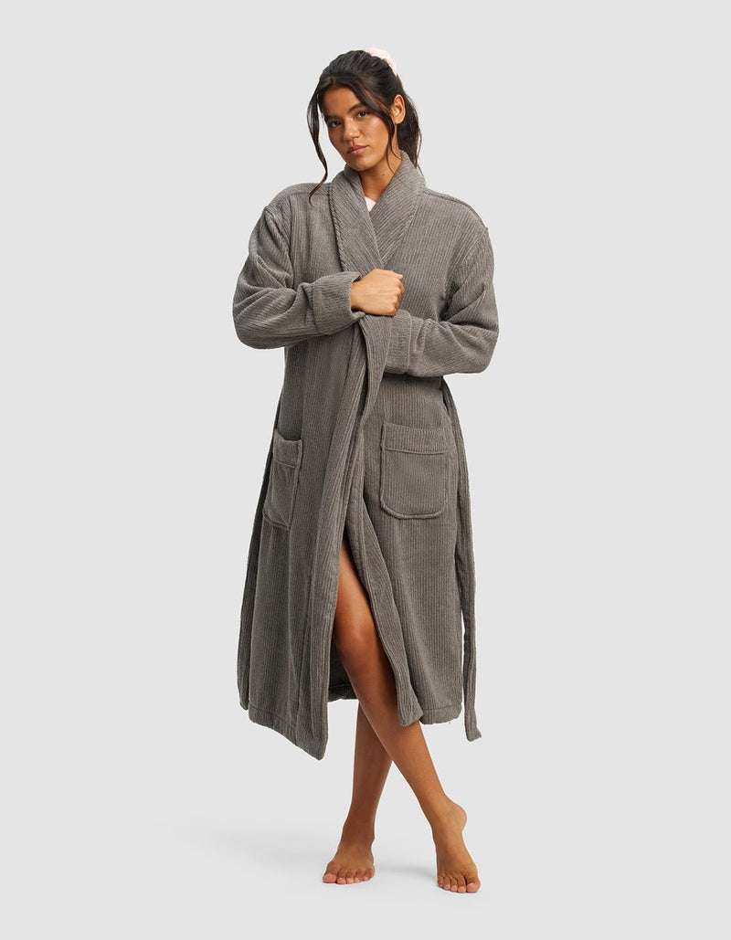 A woman with dark hair pulled back stands barefoot against a plain background, wearing Cozy Earth’s Ribbed Terry Bath Robe in gray, featuring front pockets and a waist tie. She looks slightly to the side.