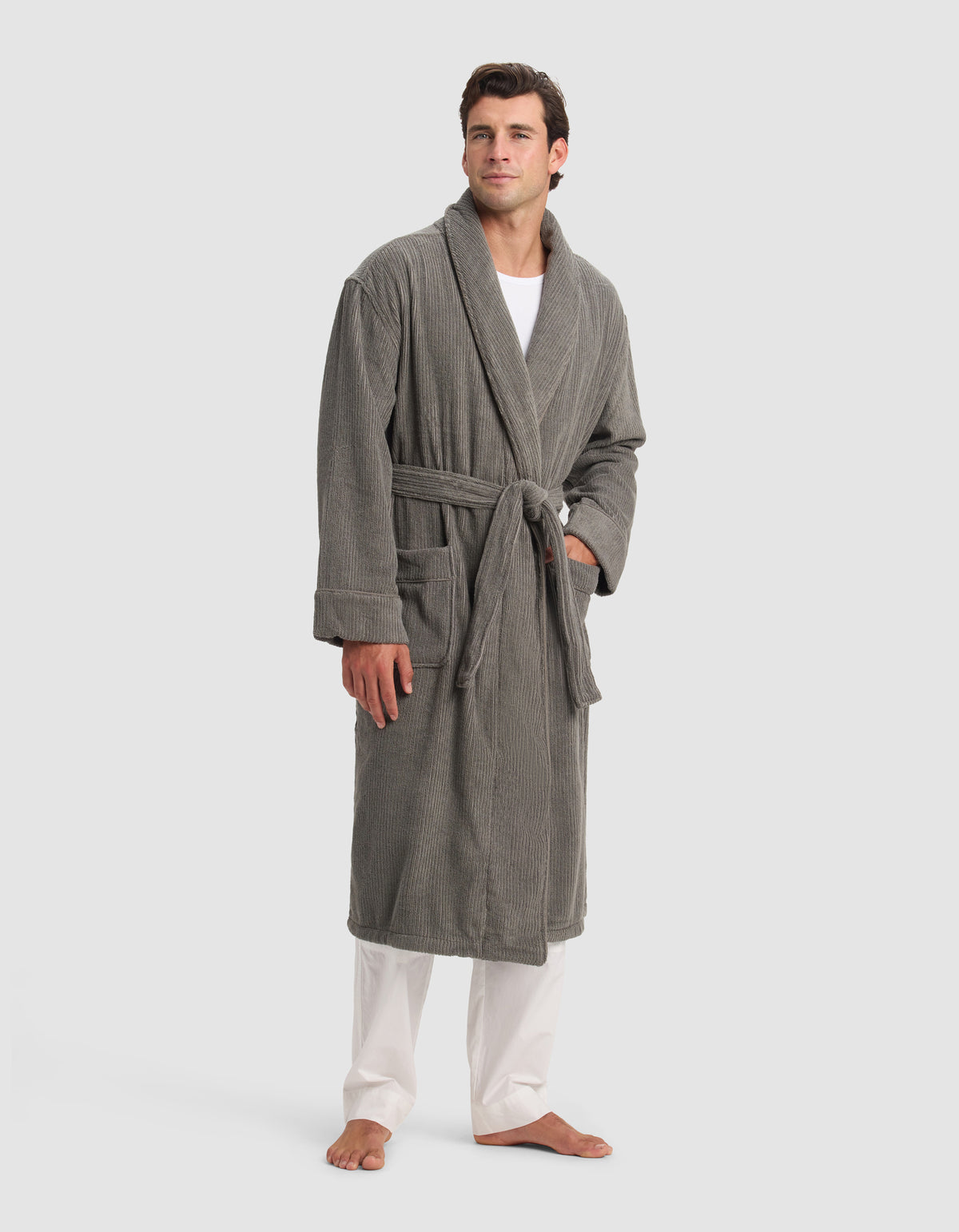 A man stands barefoot on a white background wearing the Cozy Earth Ribbed Terry Bath Robe in gray, with the belt tied at the waist. He has short brown hair and wears white pants and a white shirt underneath the robe.