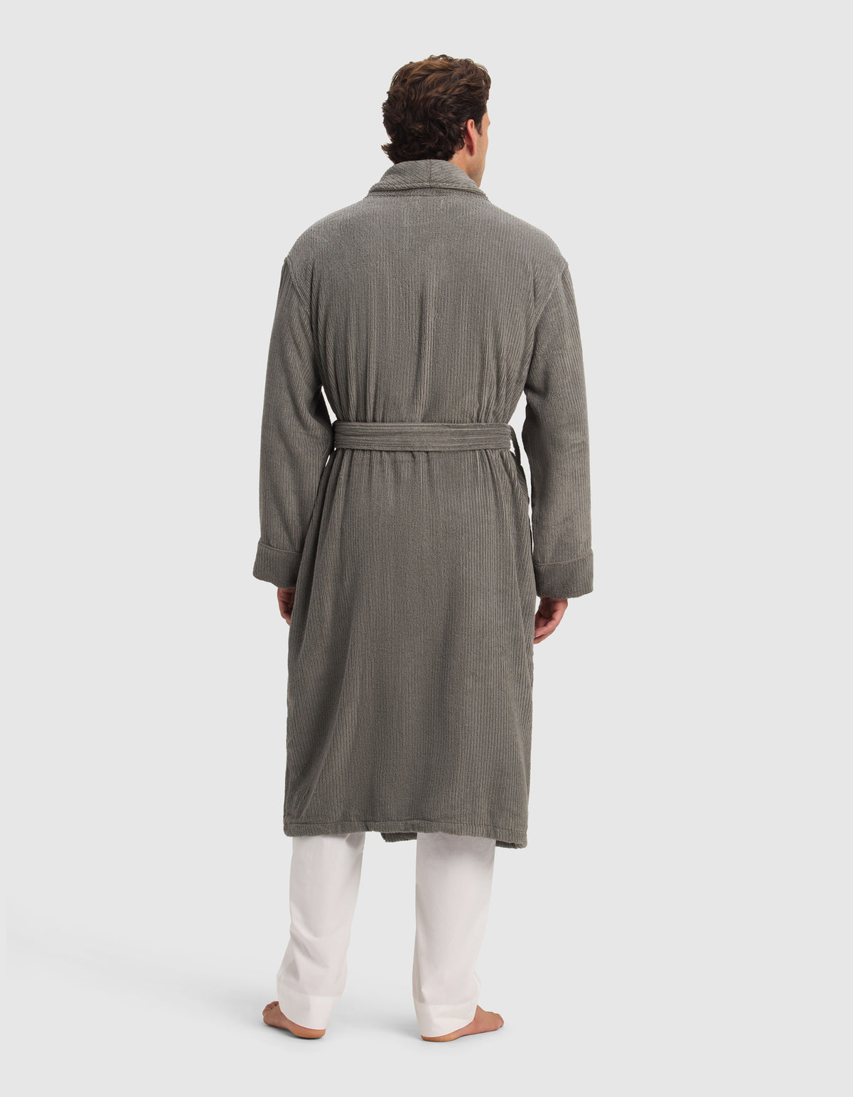 A man, barefoot against a plain light background, is wearing the Cozy Earth Ribbed Terry Bath Robe in gray, featuring a textured finish and waist belt, styled over white pants.