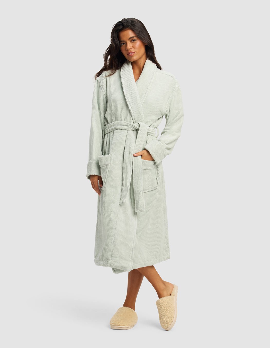 A woman with long dark hair stands against a plain background wearing the Cozy Earth Ribbed Terry Bath Robe in light sage green, featuring a belted waist and large pockets, paired with beige fuzzy slippers. She looks relaxed and comfortable.|Color:Laurel