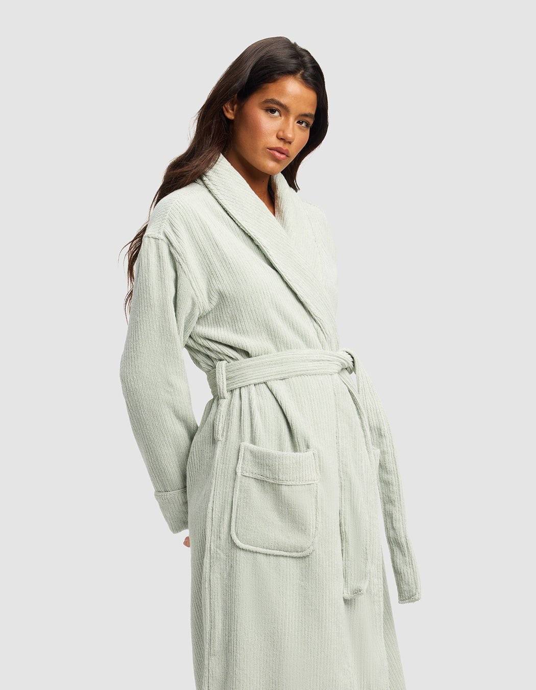 A woman with long dark hair wears the Cozy Earth Ribbed Terry Bath Robe in light sage green, featuring a belted waist and large front pockets, standing against a plain light gray background.
