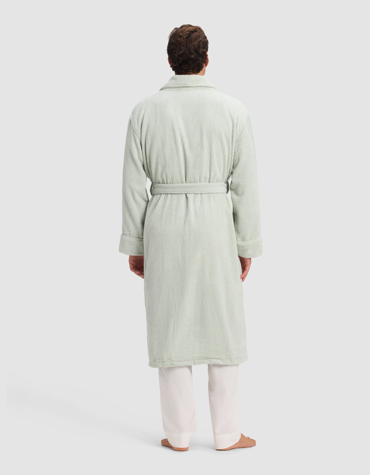 A person faces away, barefoot on a plain light background, wearing white pants and the Cozy Earth Ribbed Terry Bath Robe in light green.