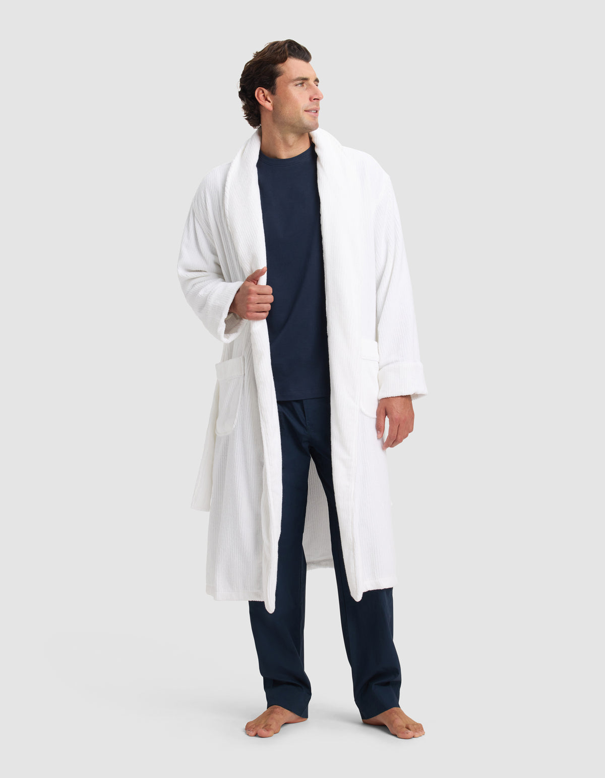A man stands barefoot in a Cozy Earth Ribbed Terry Bath Robe over a navy shirt and pants, looking left with one hand holding the robe open. The background is plain light gray.