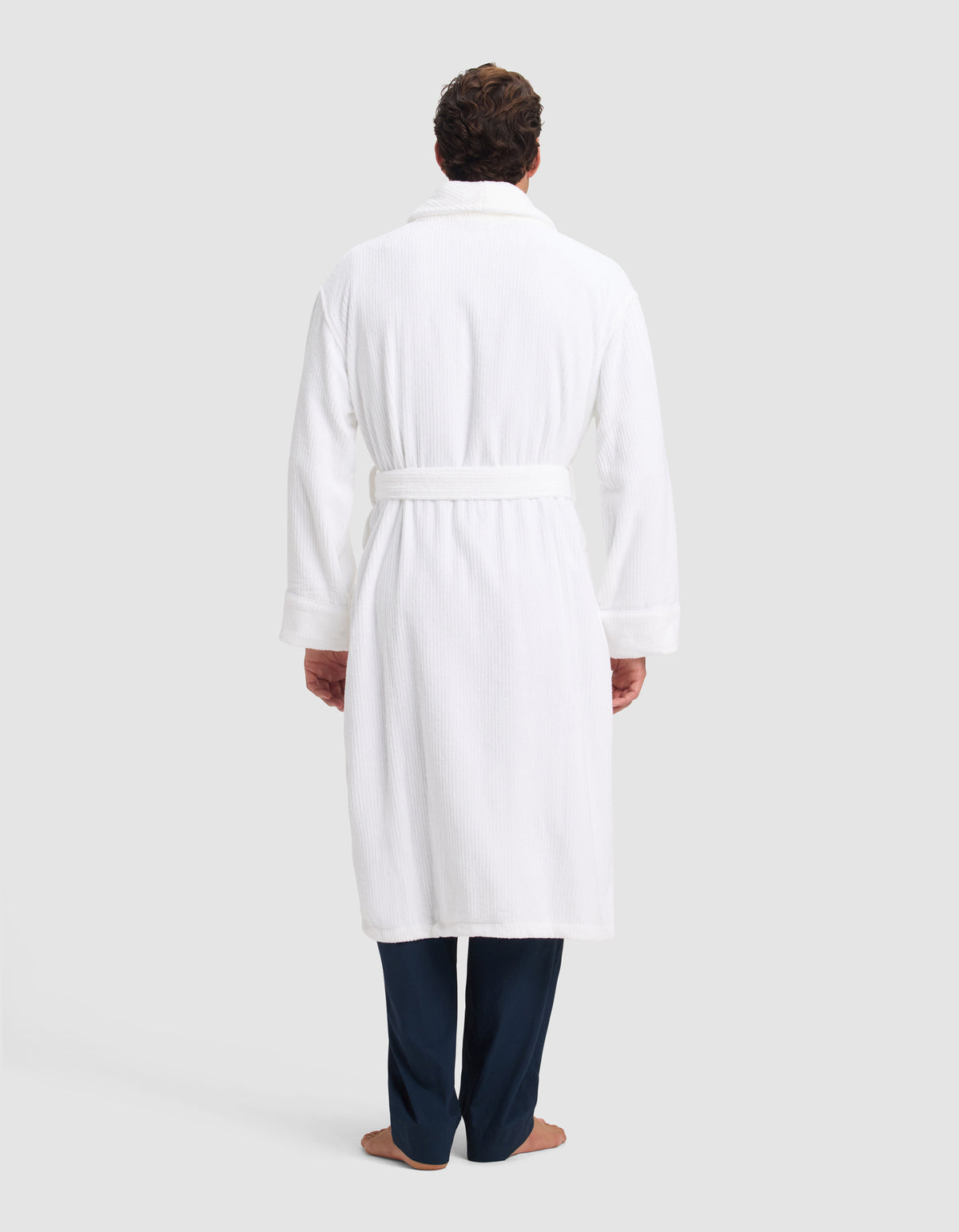 A person faces away, wearing the Cozy Earth Ribbed Terry Bath Robe with a belt, dark pants, and bare feet, set against a plain light background.