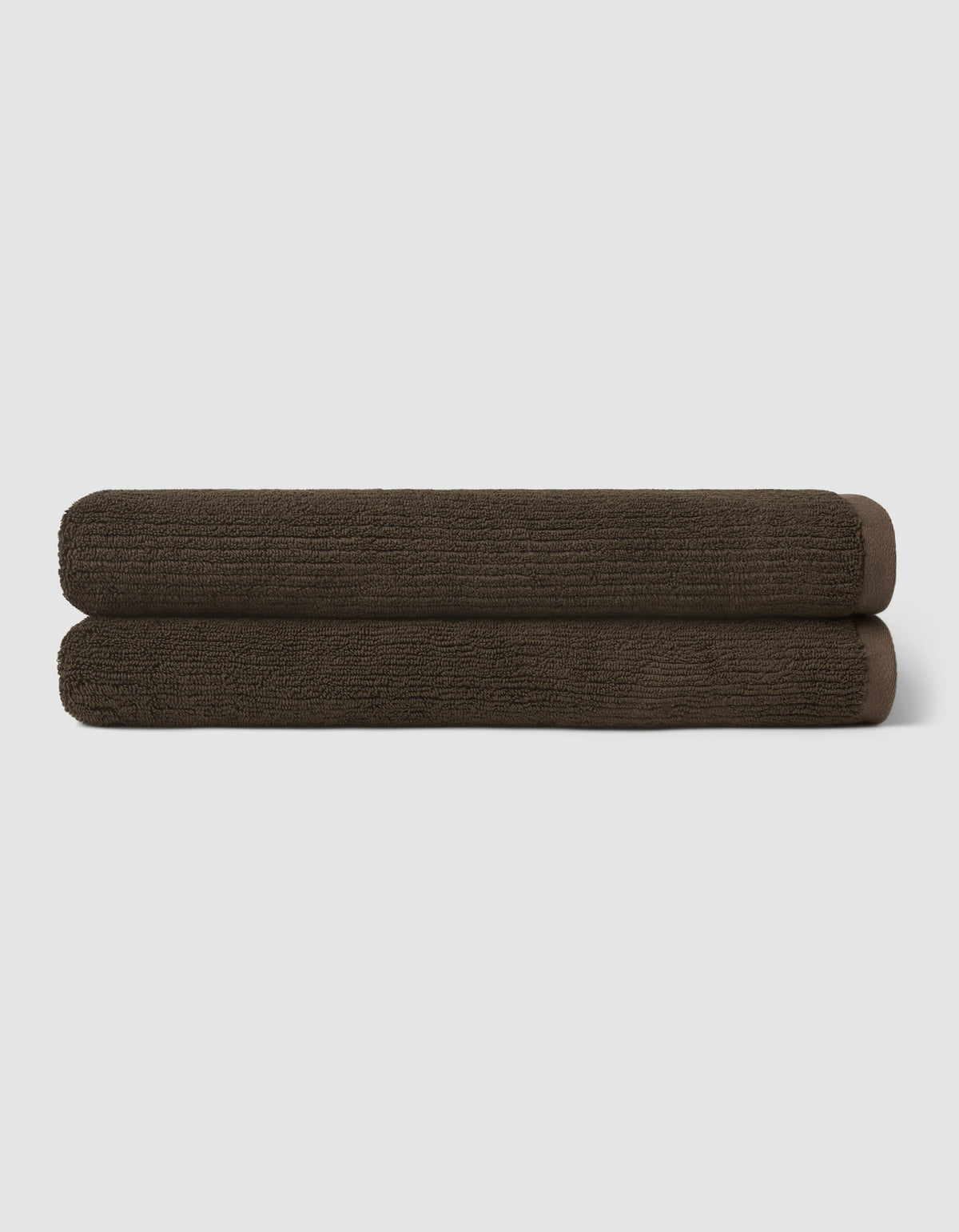 Two dark brown towels from the Cozy Earth Complete Ribbed Terry Bath Bundle are neatly rolled and stacked atop each other against a plain light gray background.|Color:Espresso