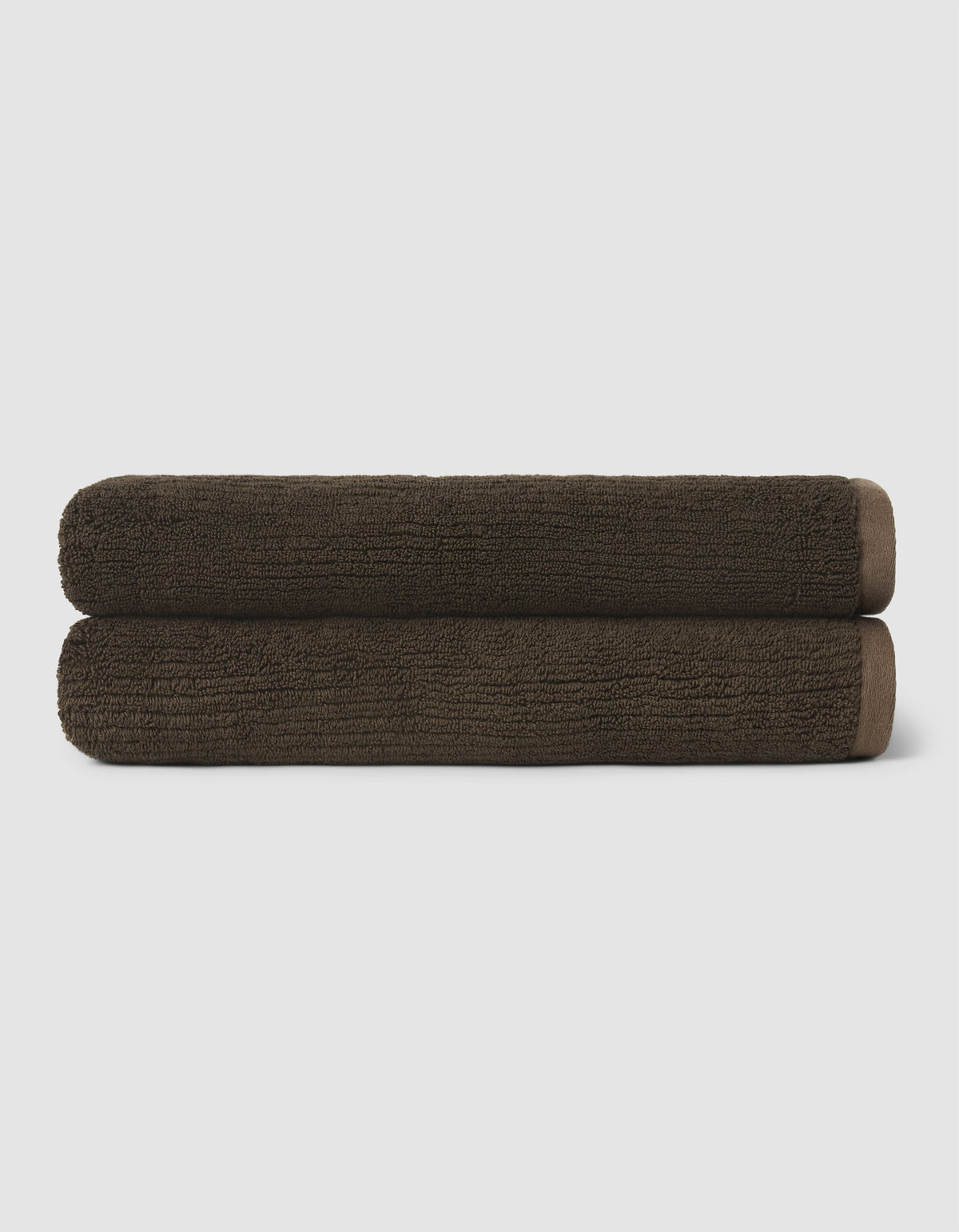 Two neatly rolled dark brown towels from the Cozy Earth Complete Ribbed Terry Bath Bundle are stacked on top of each other against a plain, light background.