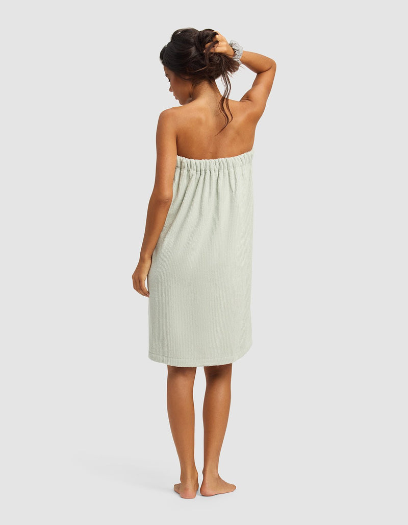 A woman stands barefoot with her back to the camera, wearing the Cozy Earth Ribbed Terry Bath Wrap in light green and a hair scrunchie, one arm raised to touch her hair, set against a plain light background.