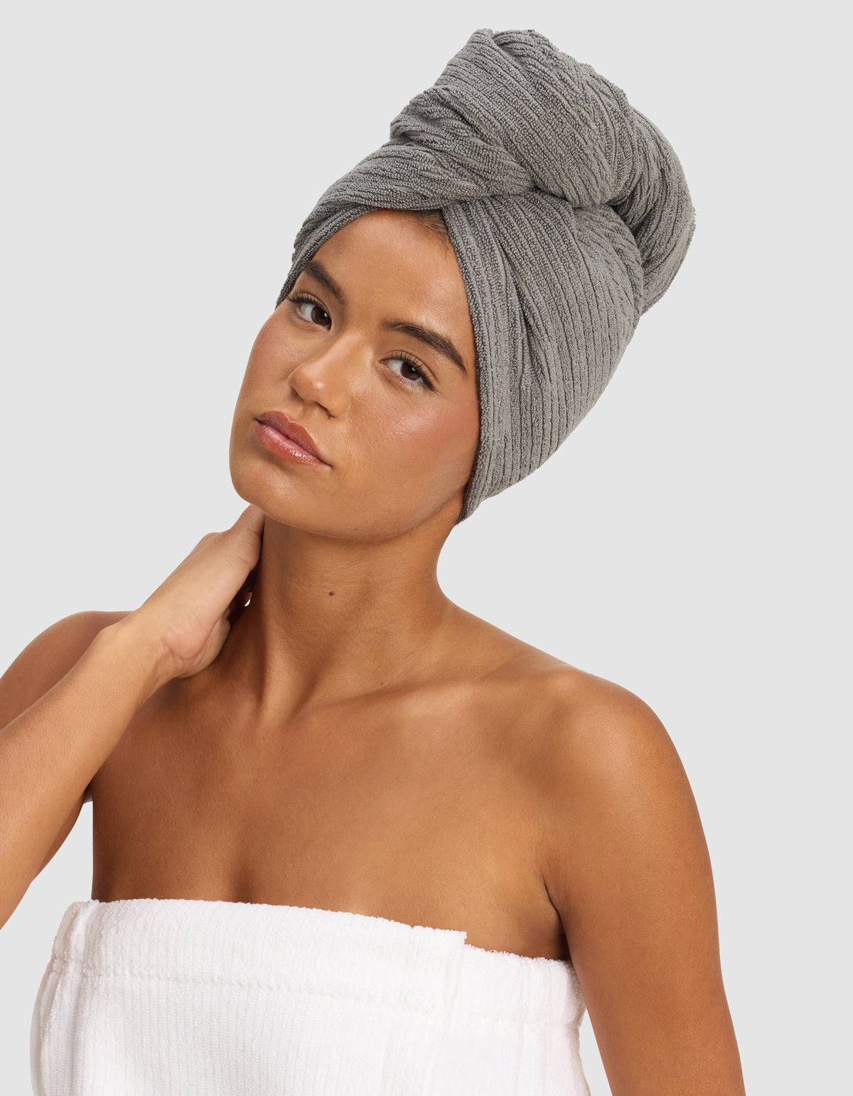 A woman poses against a plain light background, wearing the Cozy Earth Ribbed Terry Hair Towel on her head and a white towel around her body, with one hand gently touching her neck.