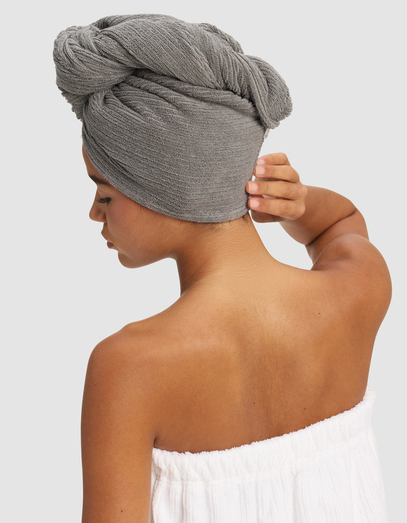 A woman with medium skin tone is seen from the back, wearing a white towel wrap and a Cozy Earth Ribbed Terry Hair Towel in gray, one hand touching the towel on her head, against a plain light background.