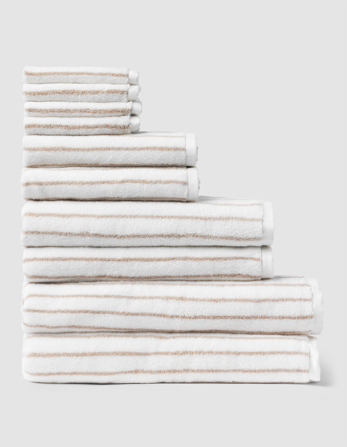 The Complete Striped Luxe Bath Bundle by Cozy Earth features neatly stacked white towels with thin beige stripes, arranged in descending size order against a plain light gray background. 