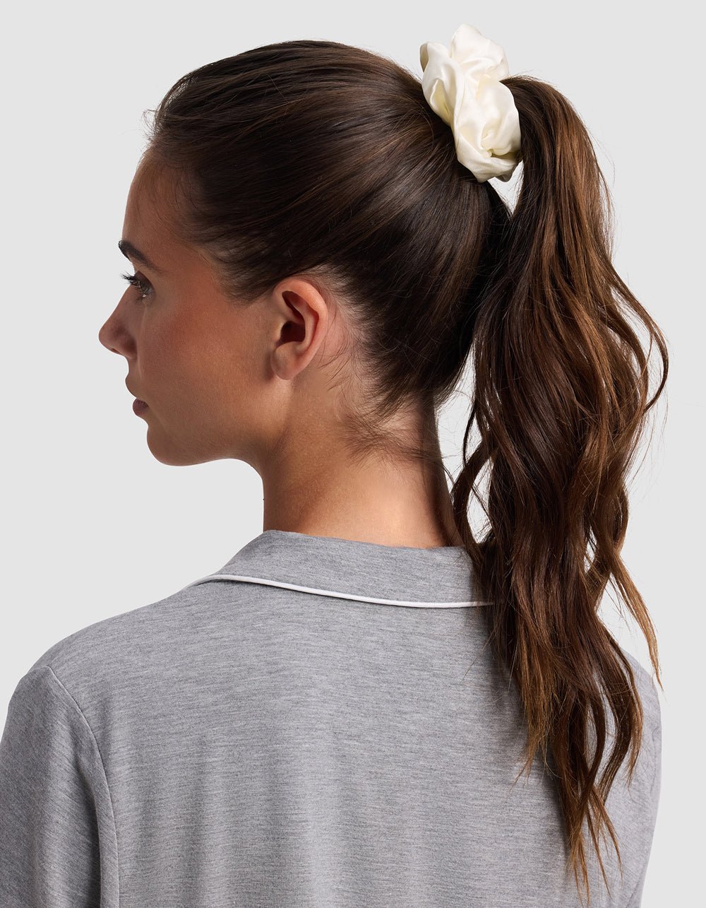A woman with long brown hair in a high ponytail, secured with a white Cozy Earth Silk Scrunchie – Last Chance, is shown from the side and back. She wears a grey top with white piping on the collar against a plain light background.
