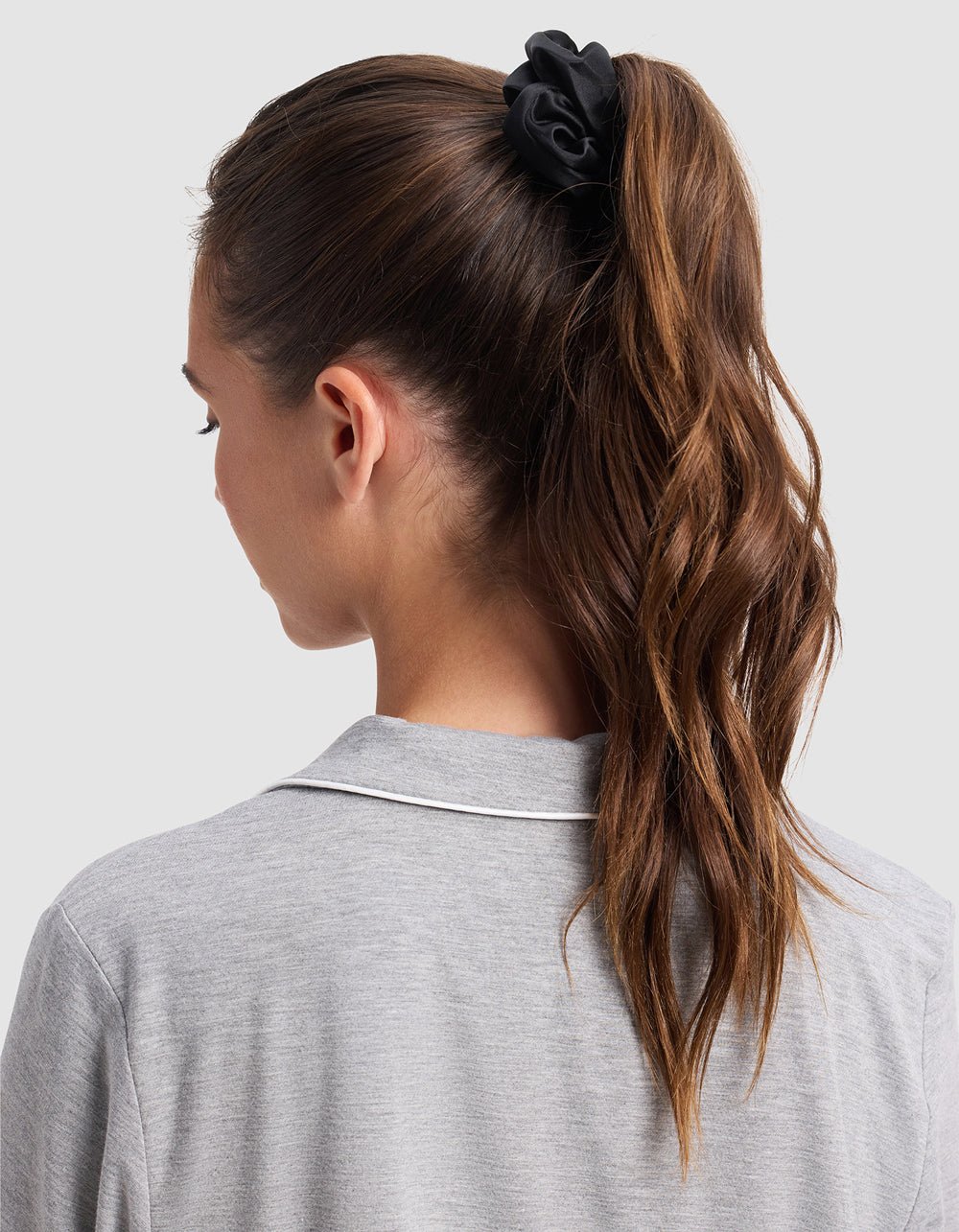 A woman with long, wavy brown hair tied in a high ponytail with a Cozy Earth Silk Scrunchie (Last Chance), wearing a light gray top with white trim, is seen from the back against a plain background.