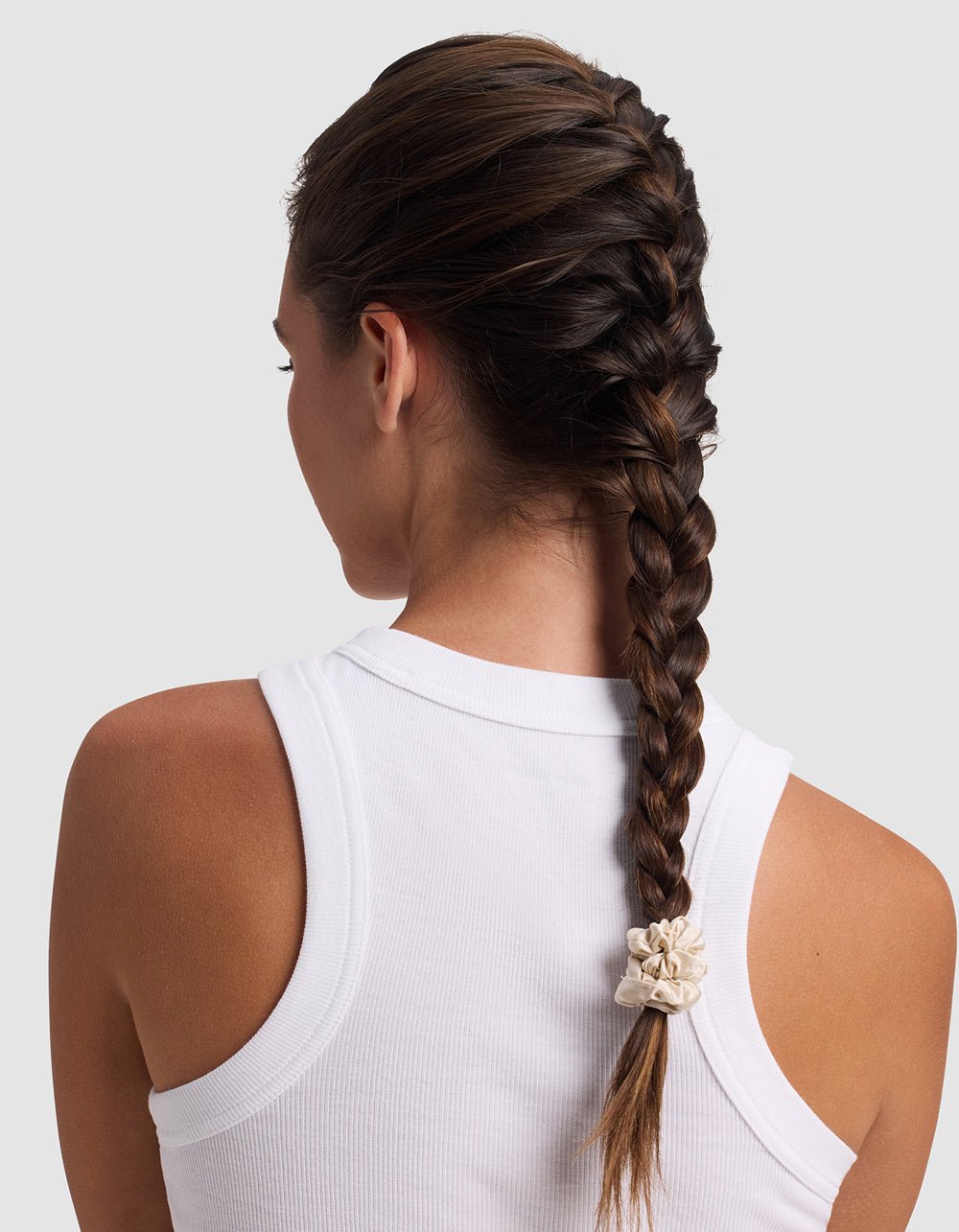 A woman with long brown hair in a French braid, secured with a Cozy Earth Silk Skinny Scrunchie, is seen from behind wearing a sleeveless white ribbed top against a plain light background.