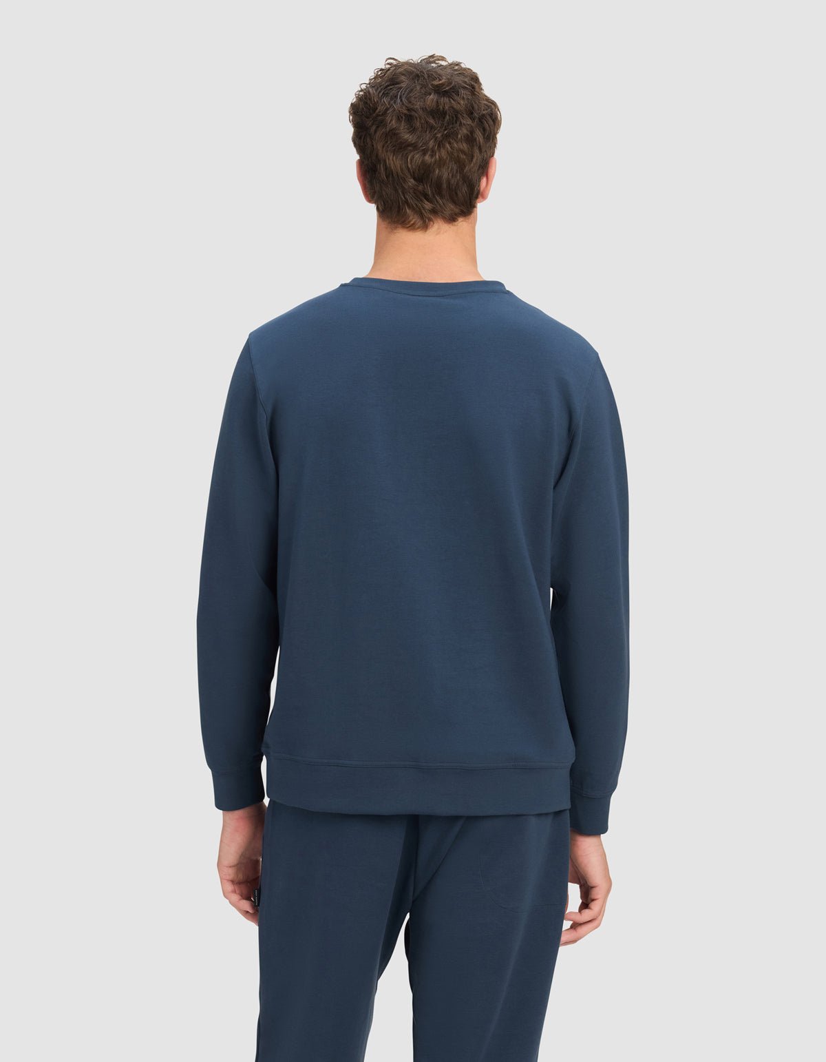 A person with short curly brown hair faces away from the camera, wearing the Cozy Earth Men's StretchTech Crewneck in dark blue against a plain light gray background.