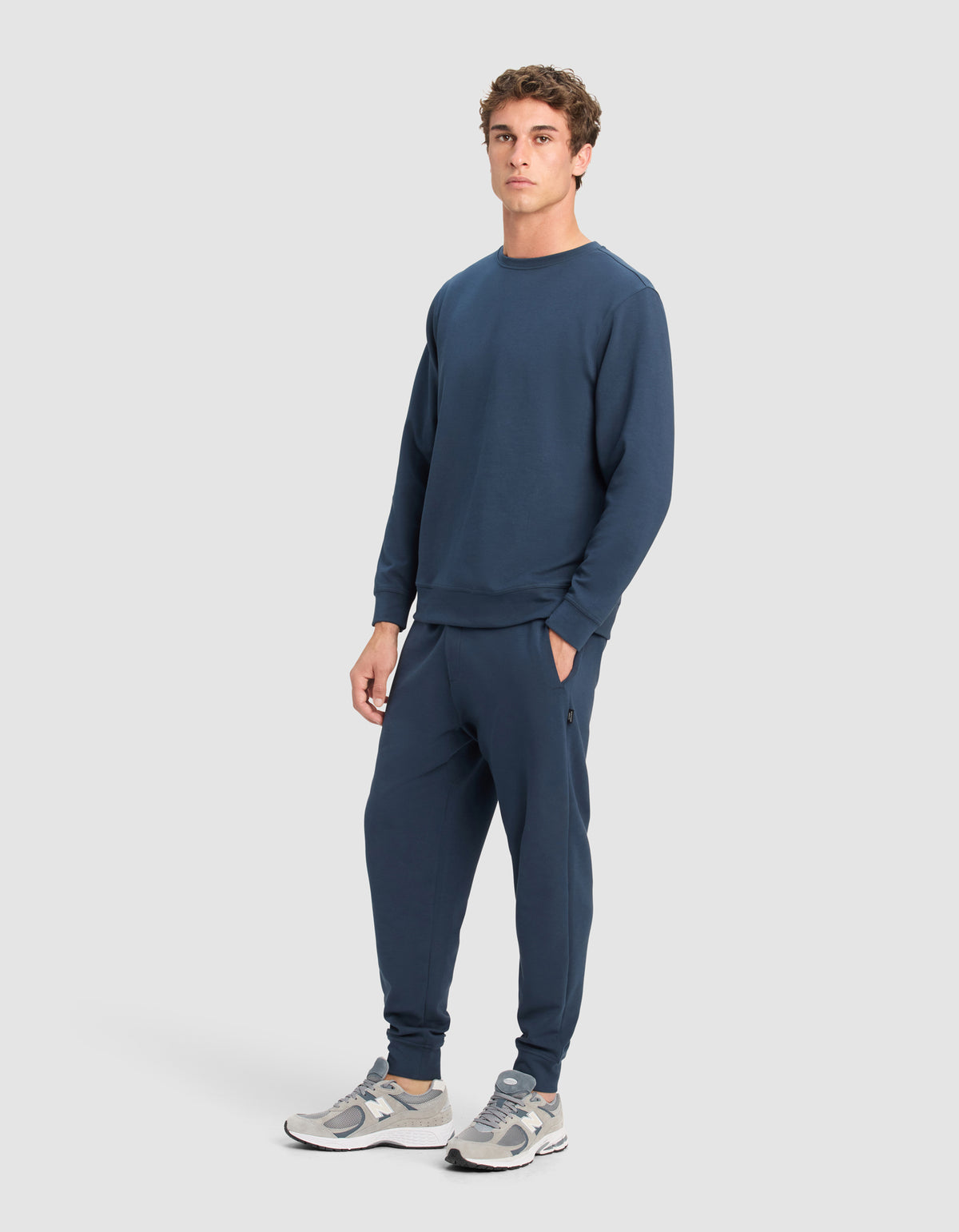 A man models the Cozy Earth Men's StretchTech Crewneck & Jogger Set in navy blue, paired with gray and white sneakers, standing facing forward with one hand in his pocket against a light gray background.
