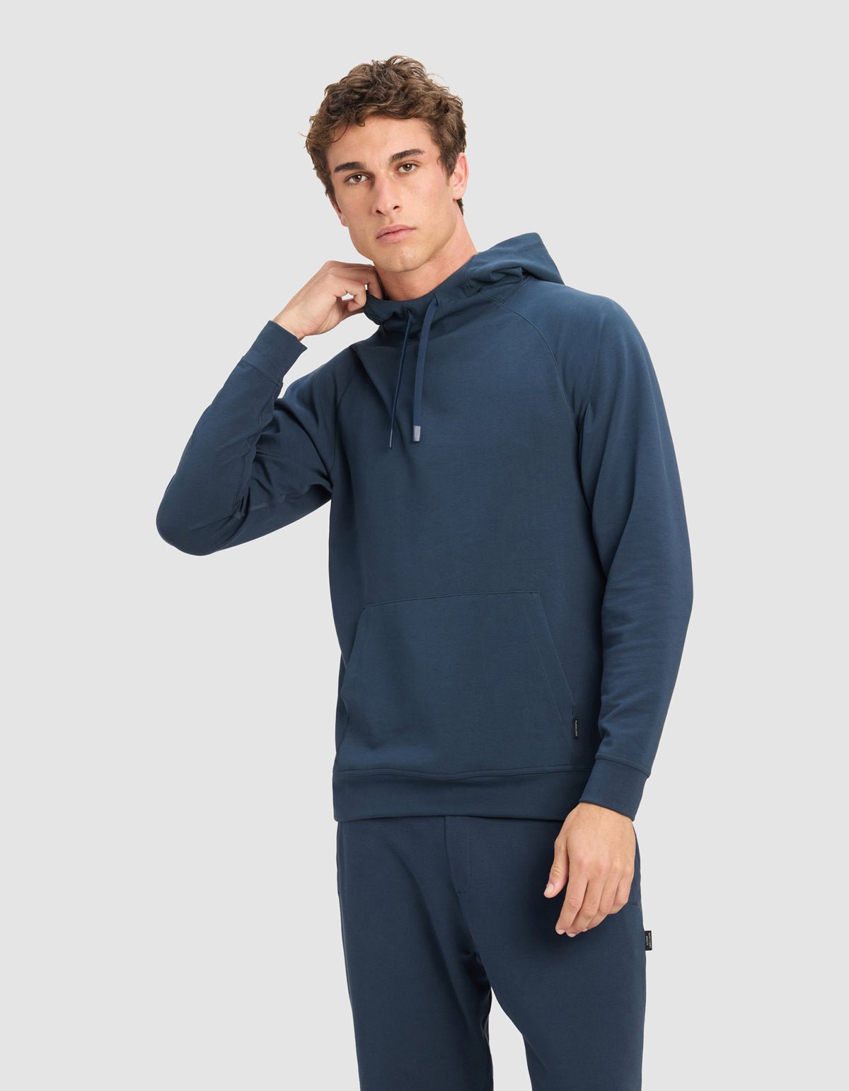 A young man models Cozy Earth's Men's StretchTech Hoodie in dark blue with matching pants, standing against a light gray background, one hand near his neck and the other by his side.