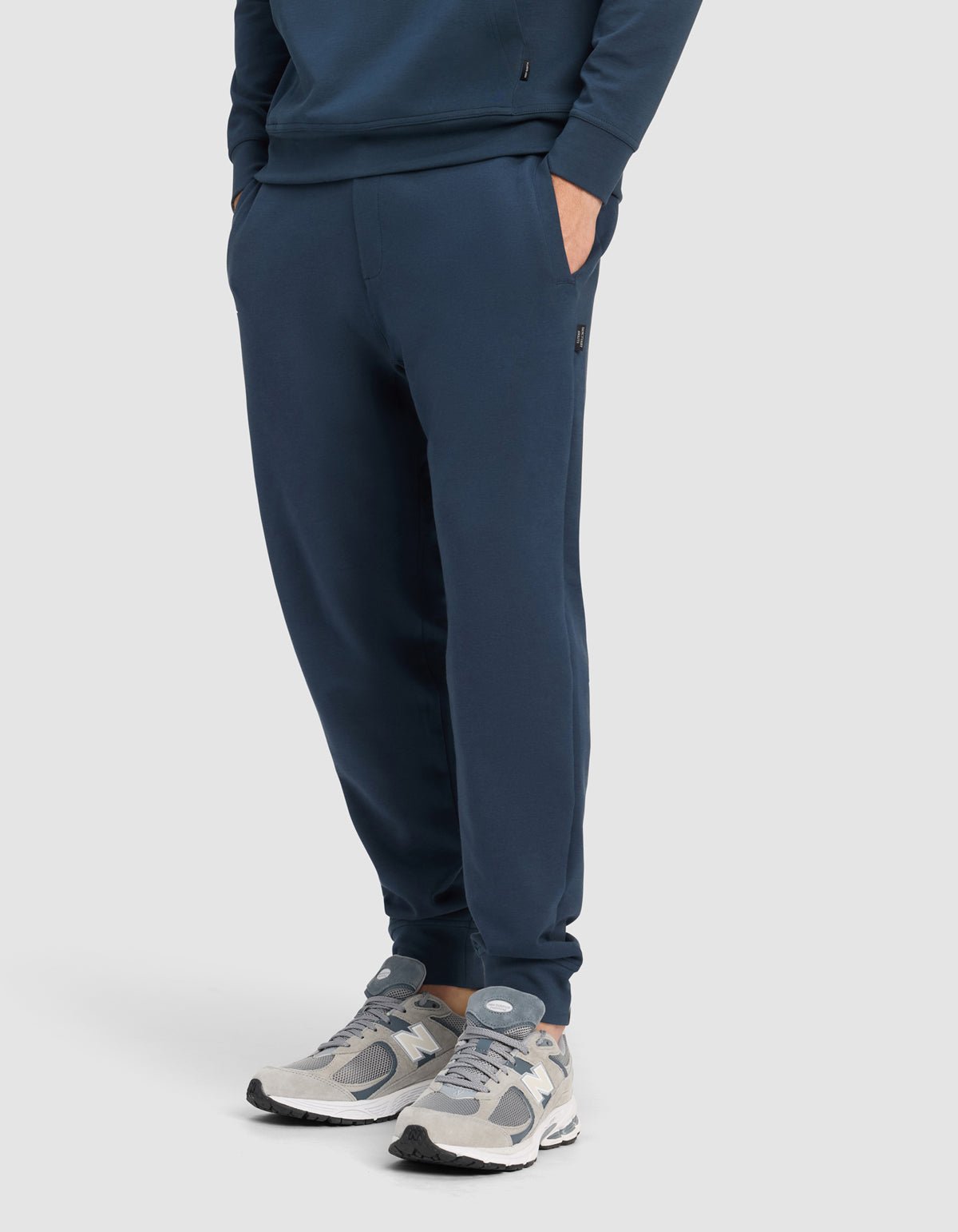 A man wears the Cozy Earth Men's StretchTech Quarter Zip & Jogger Set in navy blue, with hands in pockets, paired with gray and white sneakers, standing against a plain light background.