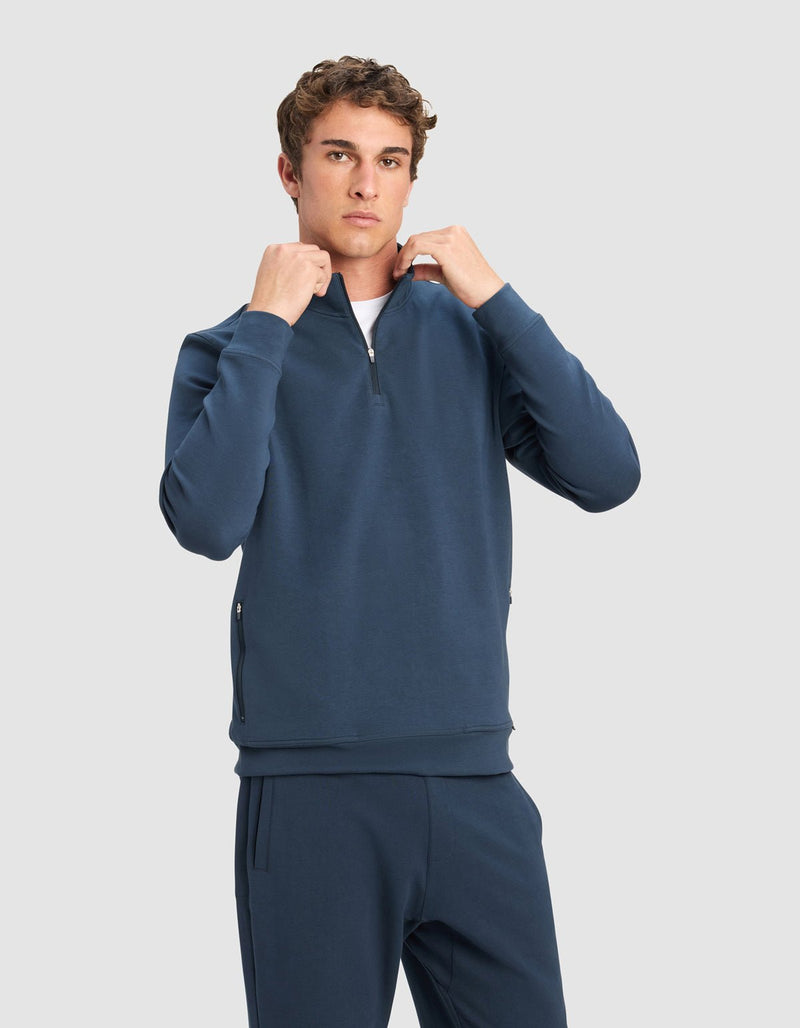 A young man with short curly hair wears the Cozy Earth Men's StretchTech Quarter Zip and matching joggers, standing against a plain light gray background and adjusting the zipper with both hands.