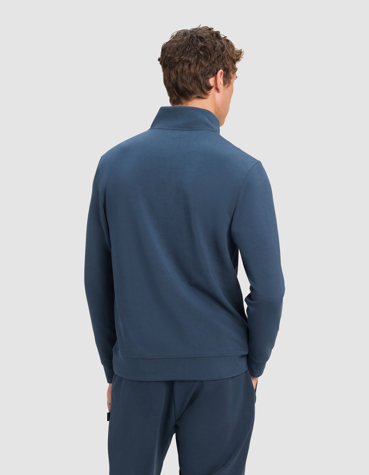 A man with curly brown hair is seen from behind wearing the Cozy Earth Men's StretchTech Quarter Zip in blue with matching pants, standing against a plain light gray background.
