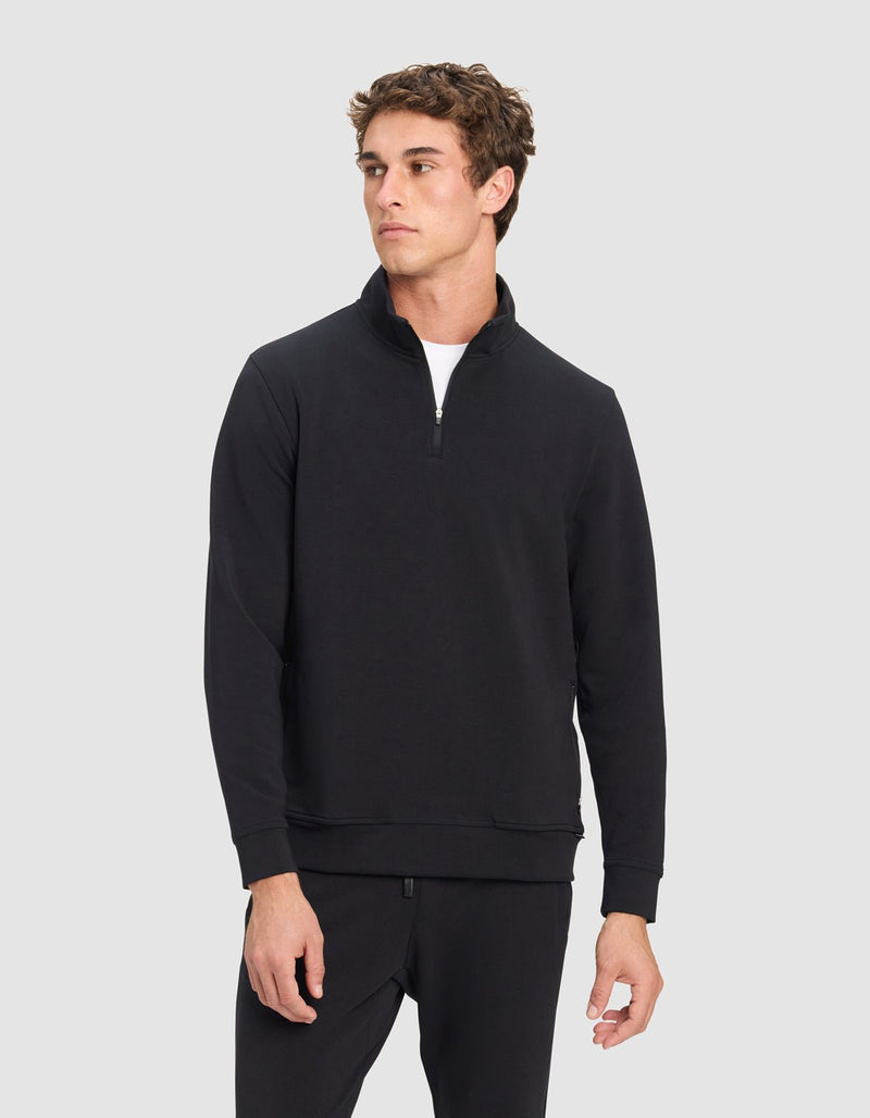 A young man in Cozy Earth's Men's StretchTech Quarter Zip and matching black pants stands against a light gray background, looking slightly to the side with a neutral expression.