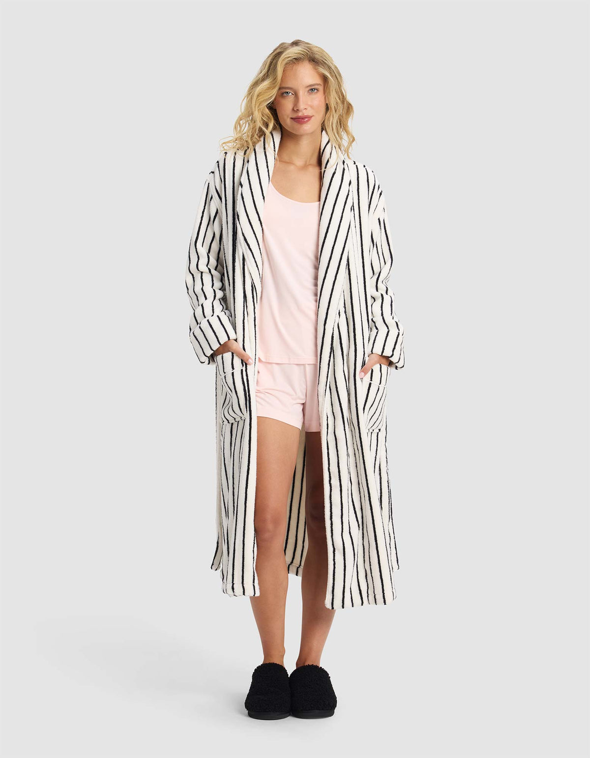 A woman stands facing forward in the Striped Luxe Bath Robe by Cozy Earth over light pink pajamas and black slippers, against a plain gray background.
