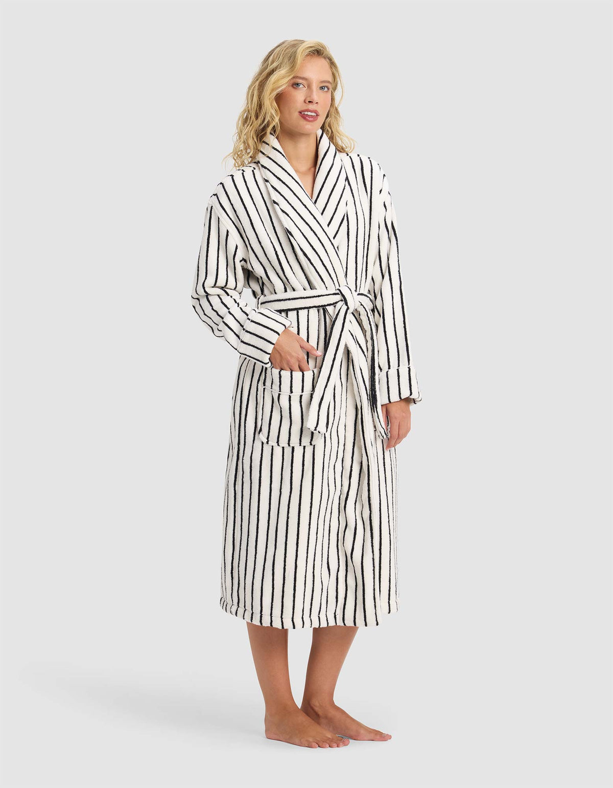A person stands barefoot in the Cozy Earth Striped Luxe Bath Robe, white with black vertical stripes and a tie waist, hands partially in pockets, against a plain light gray background.