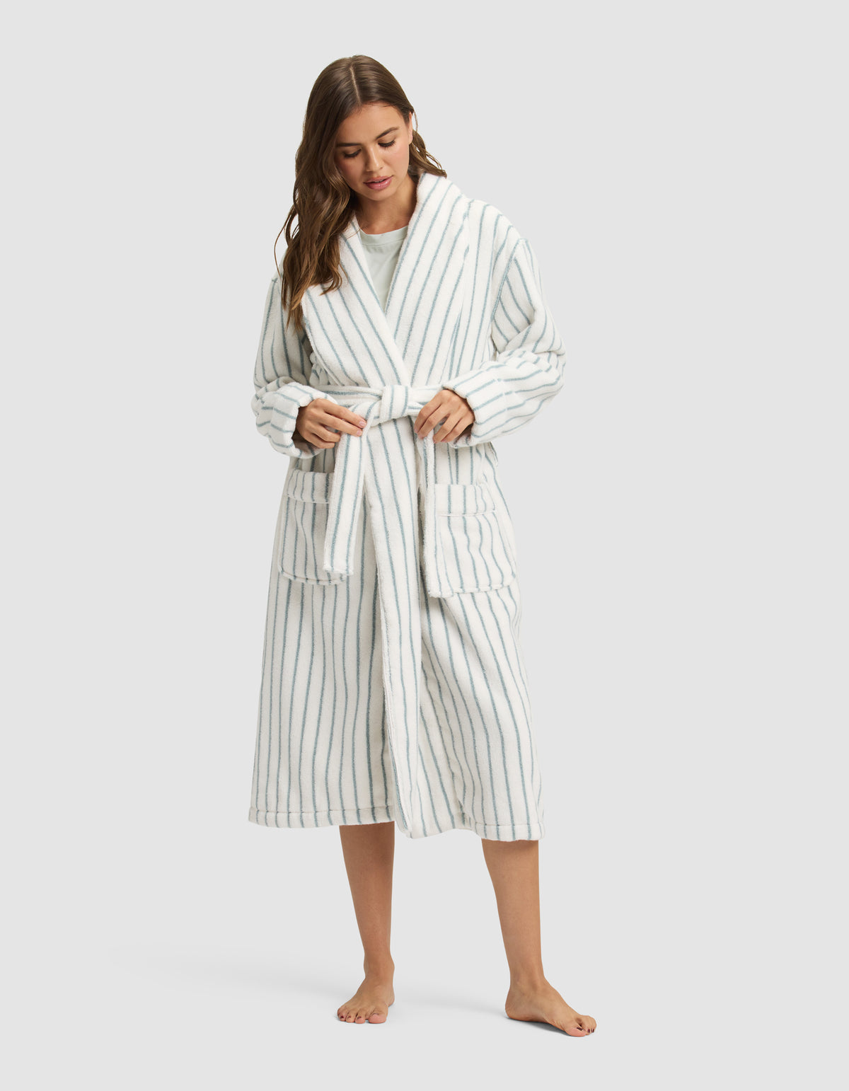 A woman, barefoot, wears the Cozy Earth Striped Luxe Bath Robe in long white fabric with vertical blue stripes, tying the waist belt as she looks downward with her long hair over one shoulder. 