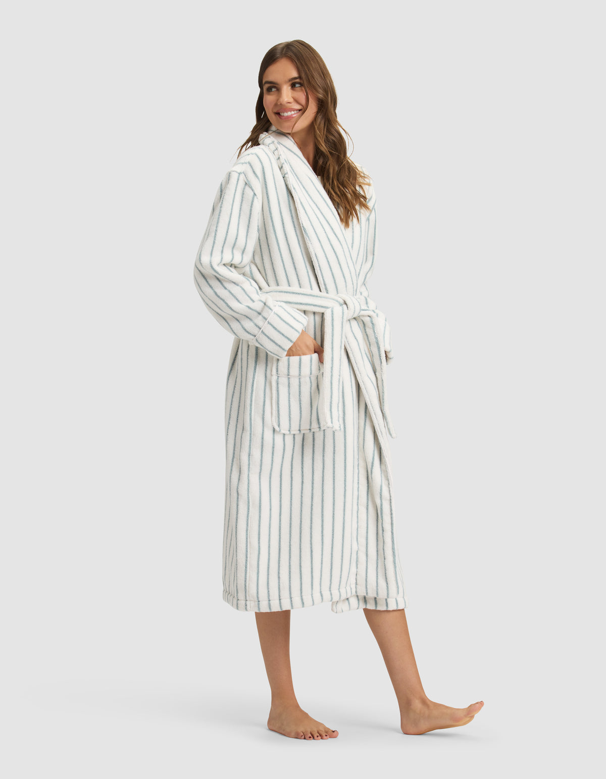 A woman stands barefoot, smiling, wearing the Cozy Earth Striped Luxe Bath Robe—long, white with blue vertical stripes, a matching belt at the waist, and large front pockets where she rests her hands. 