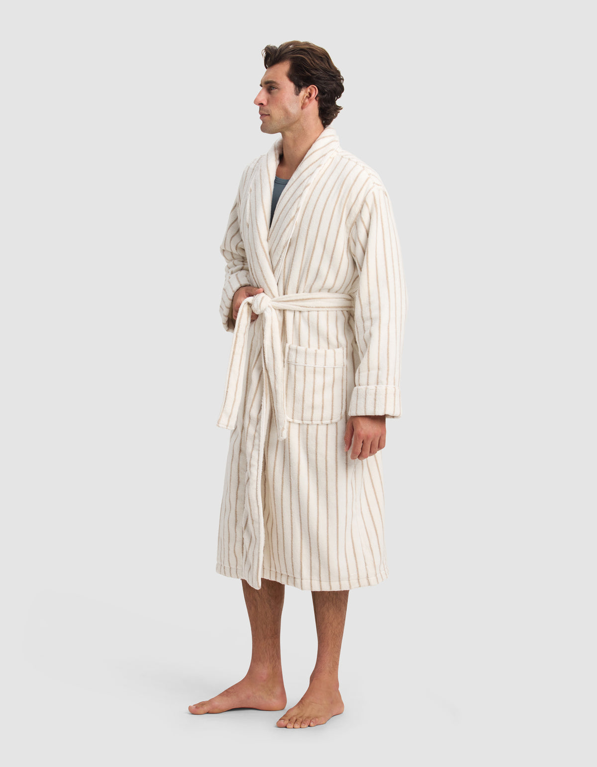 A man stands barefoot against a plain light gray background, wearing the Cozy Earth Luxe Bath Robe—a cream-colored, mid-length robe with thin vertical beige stripes and a tied belt.