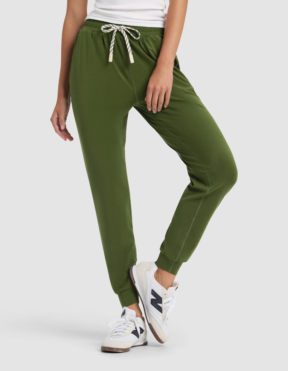 A woman stands against a plain light background wearing a white top, Cozy Earth Women's Studio Jogger pants in olive green with a drawstring, and white New Balance sneakers, one leg slightly bent.