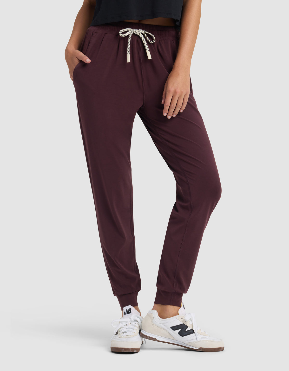 A person stands with hands in pockets against a plain light background, wearing Cozy Earth Women's Studio Jogger pants in maroon, a black top, and white New Balance sneakers.